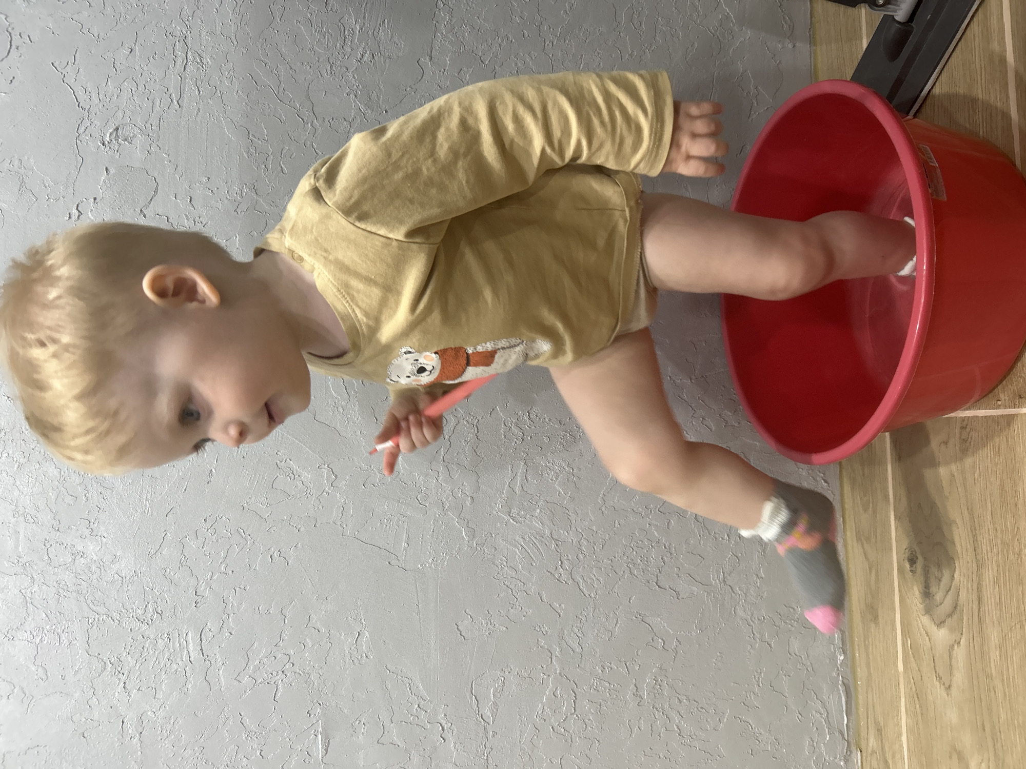 A young child with fair hair, wearing a yellow long-sleeved shirt with a bear graphic, has one leg inside a red plastic bucket. The child is holding a pink object in their hand and is looking down. The child has a gray sock with a pink toe and white ruffle at the ankle on the visible foot outside the bucket. The background shows a gray textured wall and a wood-paneled wall. A mop is visible at the upper-right corner. The location is Chaikovsky, Russia.