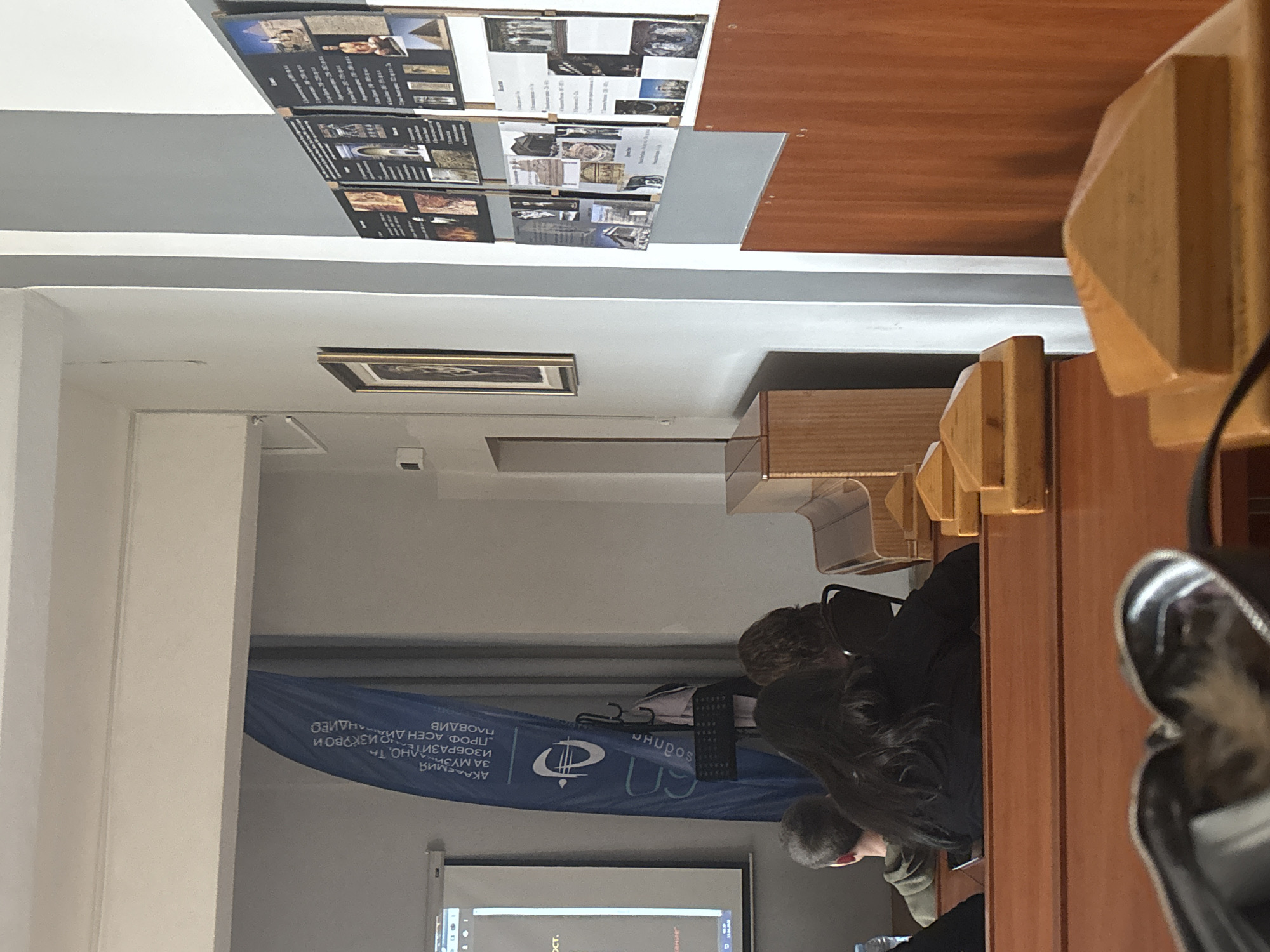Here is a detailed description of the image:
**Scene Overview:** The image appears to be taken inside a lecture hall or classroom, likely in Plovdiv, Bulgaria, based on the visible banner mentioning "Plovdiv" and "Professor Asen Diamandiev."
**People:** Several students or audience members are present. At least three individuals are partially visible, seated at desks.
**Objects:**
- Desks or tables in a row.
- Chairs.
- A large banner featuring text in Bulgarian and a logo.
- A projection screen showing a presentation.
- Informational posters about historical places hanging on the wall.
- A picture frame hanging on the wall.
**Environment:** The room seems to be moderately lit. The wall color is primarily white or off-white. The posters on the wall suggest a historical or educational context for the setting.
**Actions/Interactions:** People are seated and appear to be listening to a presentation or lecture. The projection screen indicates an ongoing presentation.