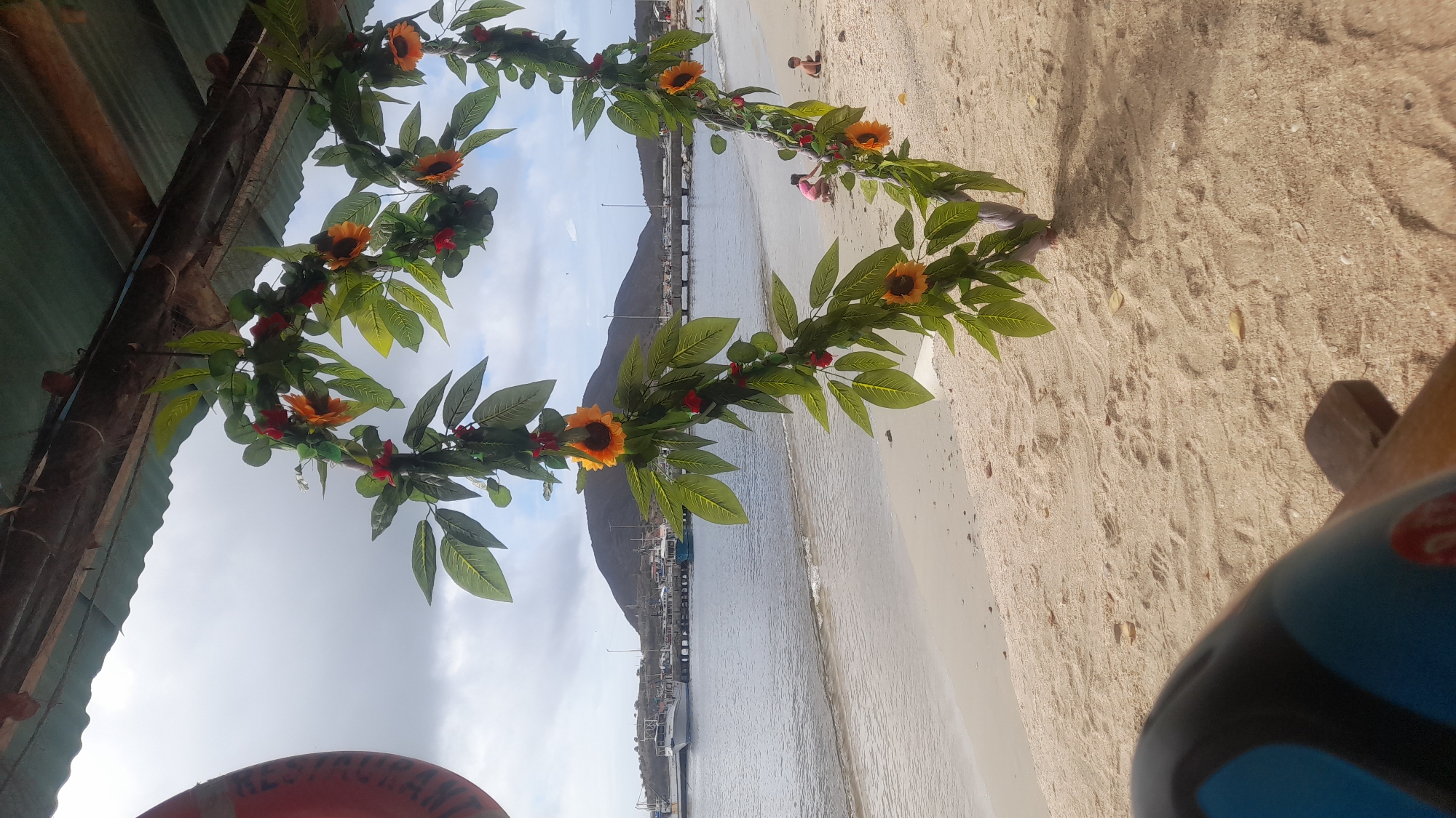 In Juan Griego, Venezuela, a heart-shaped garland of artificial sunflowers and green leaves hangs from a weathered wooden structure with a corrugated roof. The garland is positioned in the foreground, partially obscuring a view of the beach, the ocean, and a distant mountainous landscape. The beach is sandy, with some scattered leaves and footprints. Two children are visible on the sand near the water's edge, one in pink and one in a light-colored shirt. The ocean is calm, with a few small waves lapping the shore. In the background, a pier extends into the water, lined with boats. The sky is cloudy, suggesting an overcast day. The lower left corner of the image shows a blurred section of what appears to be a seat or chair, with a red and blue design.
