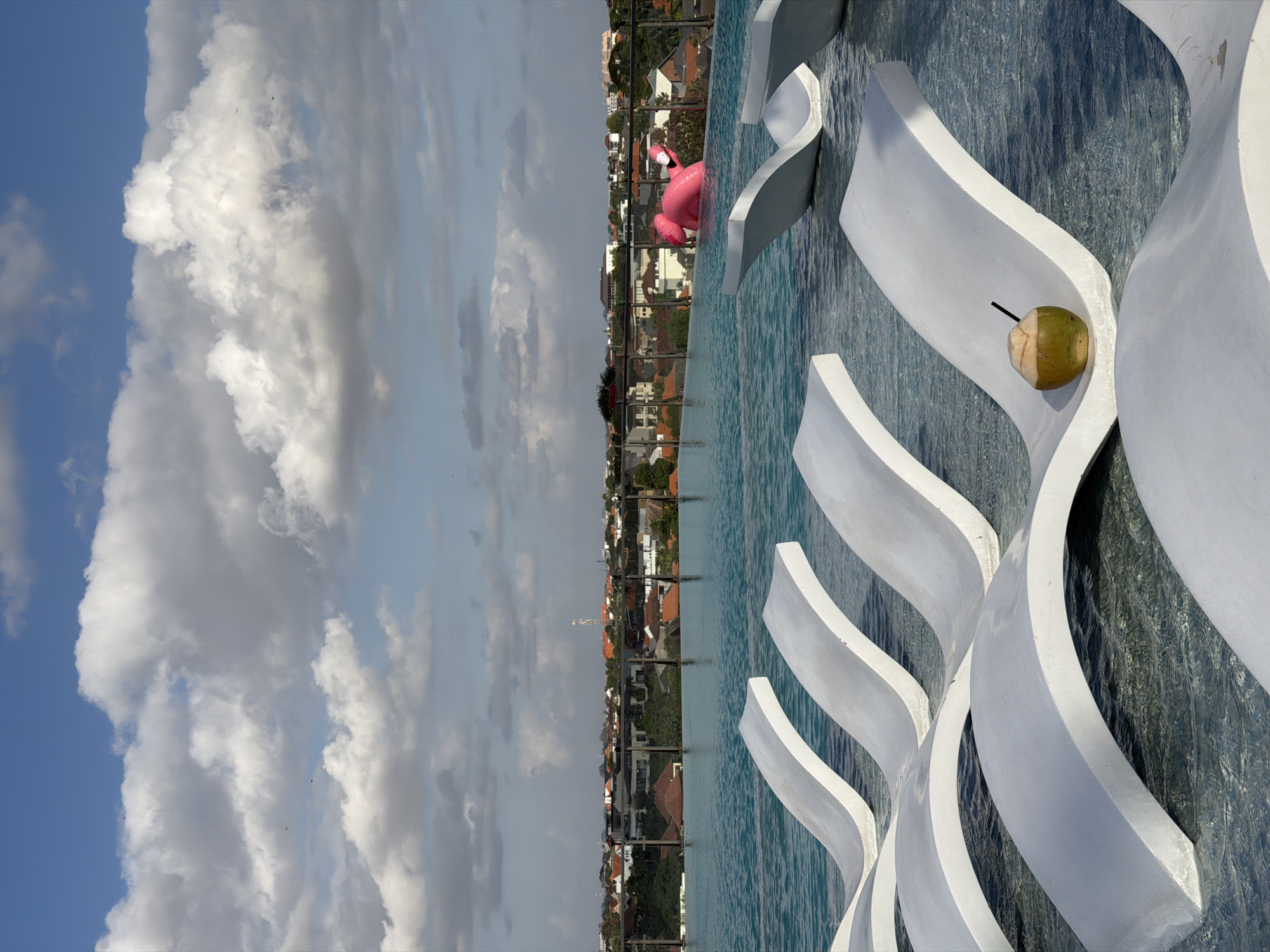 An outdoor infinity pool is shown, with white chaise lounges partially submerged in the clear blue water. A whole coconut with a black straw rests on one of the chaise lounges. In the background, a pink inflatable flamingo floats in the pool. Beyond the pool, a cityscape with red-tiled roofs and scattered greenery is visible under a bright blue sky with fluffy white clouds. The location is identified as North Kuta, Indonesia.
