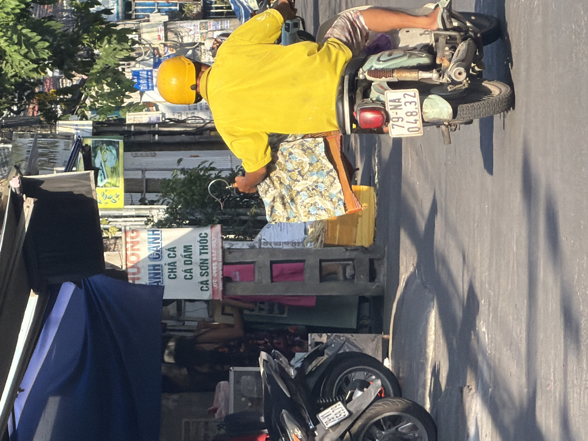 A medium shot captures a street scene in Nha Trang Ward, Vietnam, on a sunny day. In the foreground, a person wearing a bright yellow shirt and a yellow helmet rides a scooter away from the viewer. The scooter has a license plate that reads "79-NA 048.32". The rider is carrying a patterned cloth draped over the handlebars. To the left, another scooter is parked. In the background, a storefront is visible, with a blue awning and a sign in Vietnamese. A person can be seen inside the store. The environment suggests a bustling street with buildings and trees.