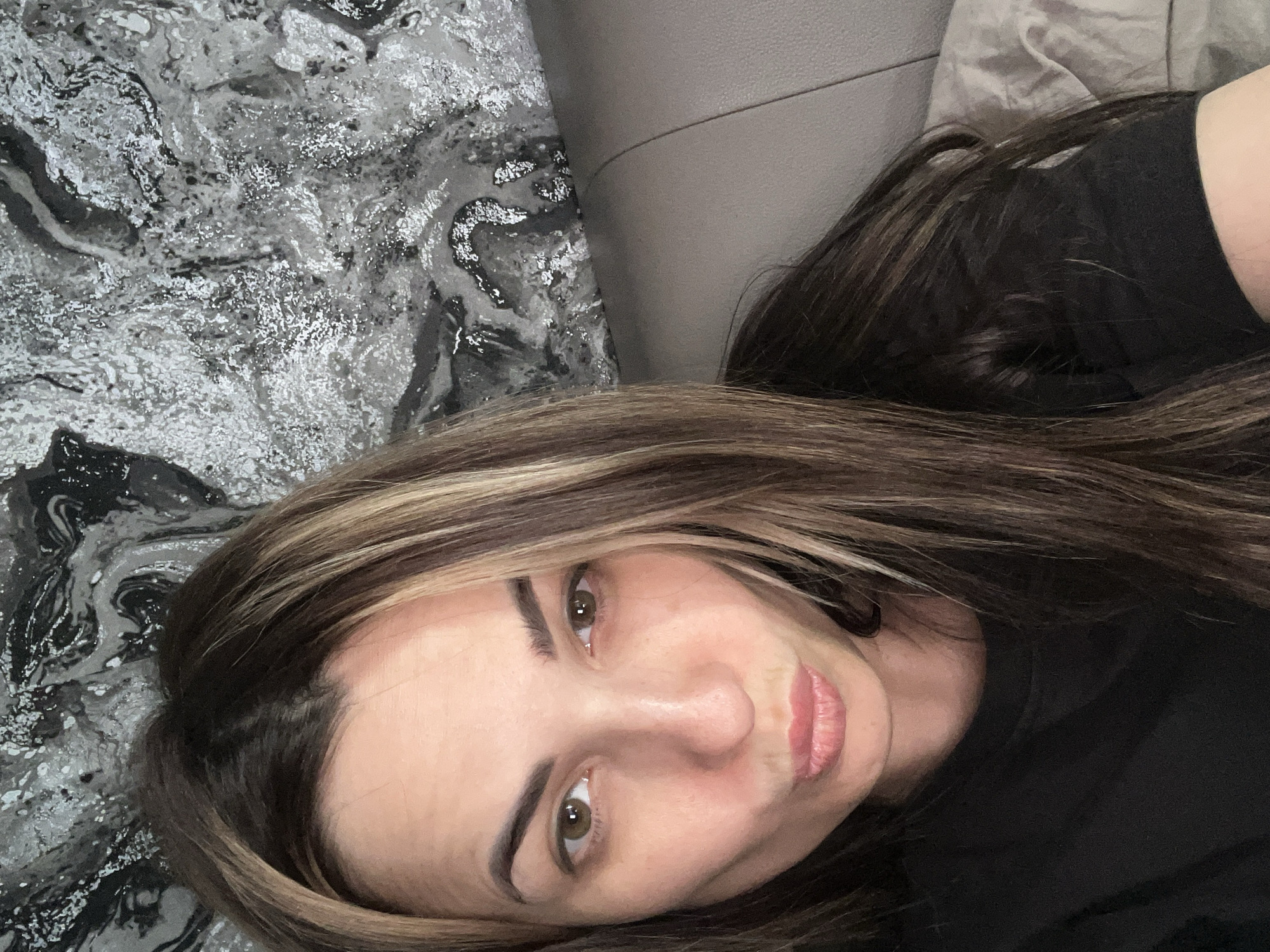 A woman with brown hair and blonde highlights is lying down, looking at the camera. She is wearing a black shirt. She has brown eyes, fair skin, and pink lips. The background includes a grey marble-patterned surface, a light grey cushion, and a darker grey area. The location is Unknown, Unknown.