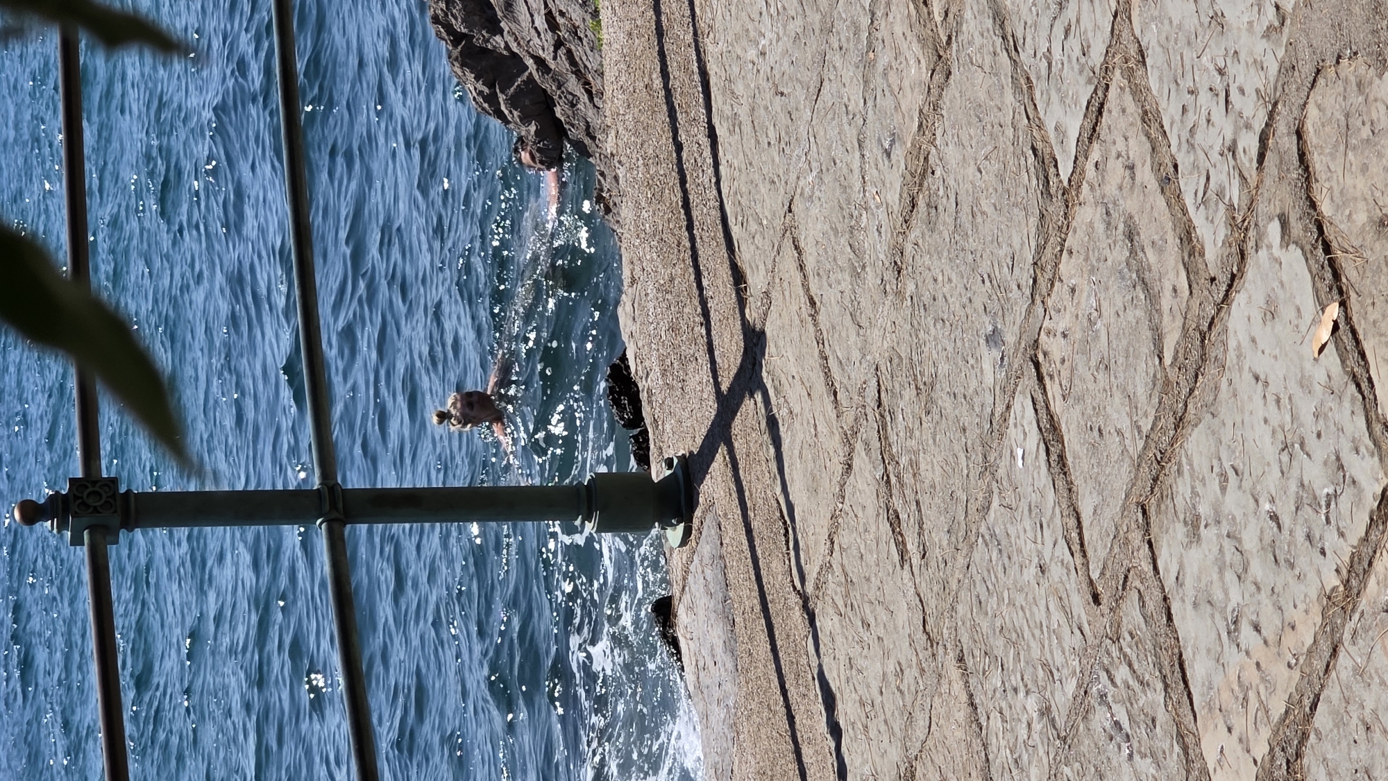 Two individuals are swimming in the Adriatic Sea near Opatija, Croatia. The water is blue and choppy, with sunlight glinting off the surface. In the foreground, a stone walkway is visible, with a metal railing and a green post casting a shadow. The walkway is paved with large, irregularly shaped stones. The sea is to the left of the walkway, and a rocky outcrop is on the right. The person on the left is a woman with her hair pulled up in a bun, swimming on her back. The person on the right is partially submerged, with only their head and shoulders visible. The overall scene suggests a sunny day at the coast.