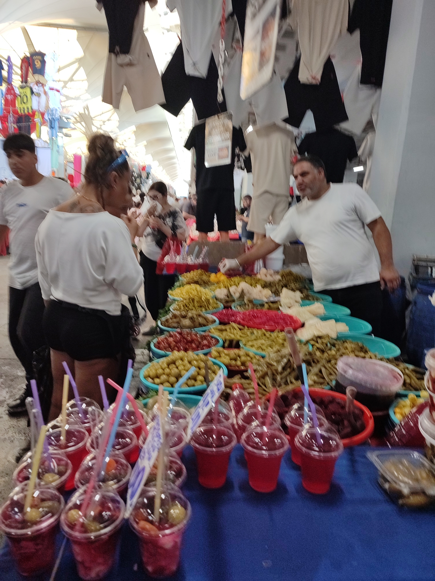 This is a street scene in Kadıköy, Turkey. A man in a white t-shirt is standing behind a market stall filled with various types of pickles and olives. He is wearing gloves and appears to be serving a customer, as his right hand is reaching towards the produce. In the foreground, there are several plastic cups filled with a red beverage and fruit, each with colorful straws. Behind the stall, two women and a man are visible, one woman is wearing a white shirt and black shorts and the other a patterned top and dark pants. The background shows a bustling market environment with clothing items hanging from above, suggesting a lively shopping area.