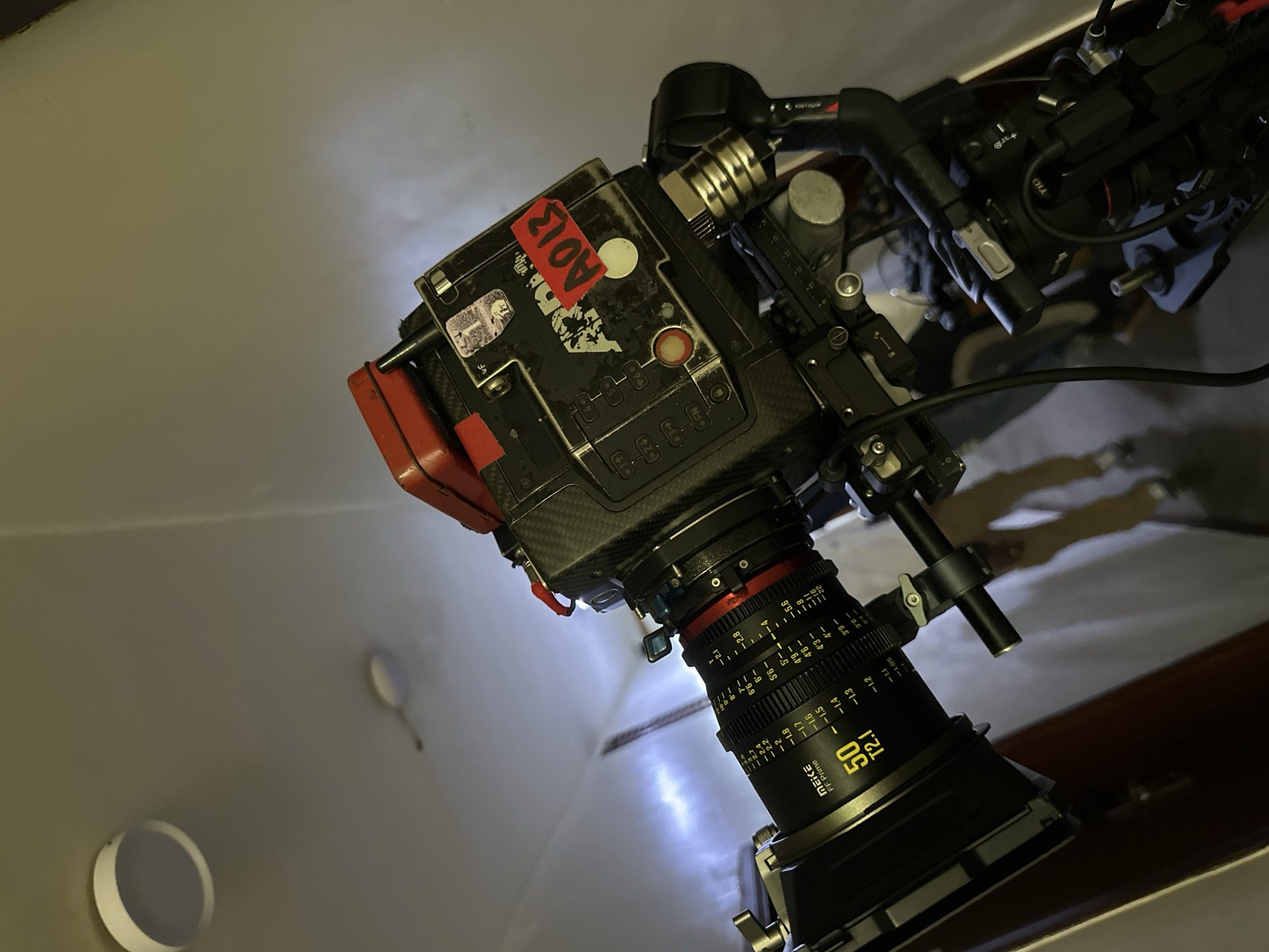 The image shows a high-end camera, possibly a cinema camera, mounted on some kind of rig. The lens is a Meike FF Prime 50mm T2.1. The camera body has various stickers and a label that reads "A013." The background appears to be a plain, possibly painted wall with some recessed lighting. The location is unknown, and there is no indication this image was taken in Lagos, Nigeria. There are no people visible.