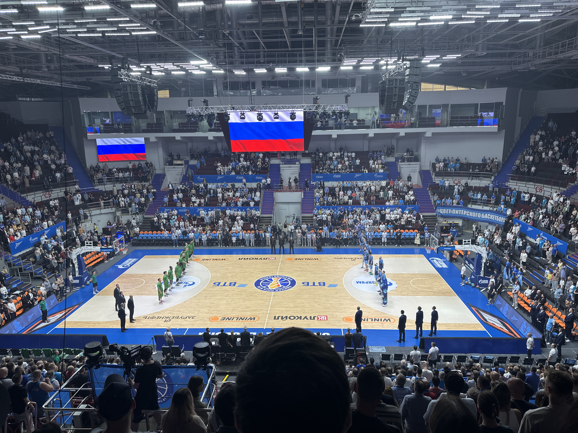 The image shows a basketball game taking place in a large indoor arena filled with spectators. Two teams are lined up on the court, one in green uniforms and the other in blue. Several officials in suits stand near the center of the court. Advertisements from various companies, including BTB and Winline, are visible on the court. The Russian flag is displayed on screens above the audience on both sides of the stadium. The scene appears to be taking place in Saint Petersburg, Russia, as a sign on the edge of the court reads "ЦВЕТ ПИТЕРСКОГО БАСКЕТБОЛА" which translates to "Saint Petersburg basketball color". A sign with "ST PETERS" is also visible.