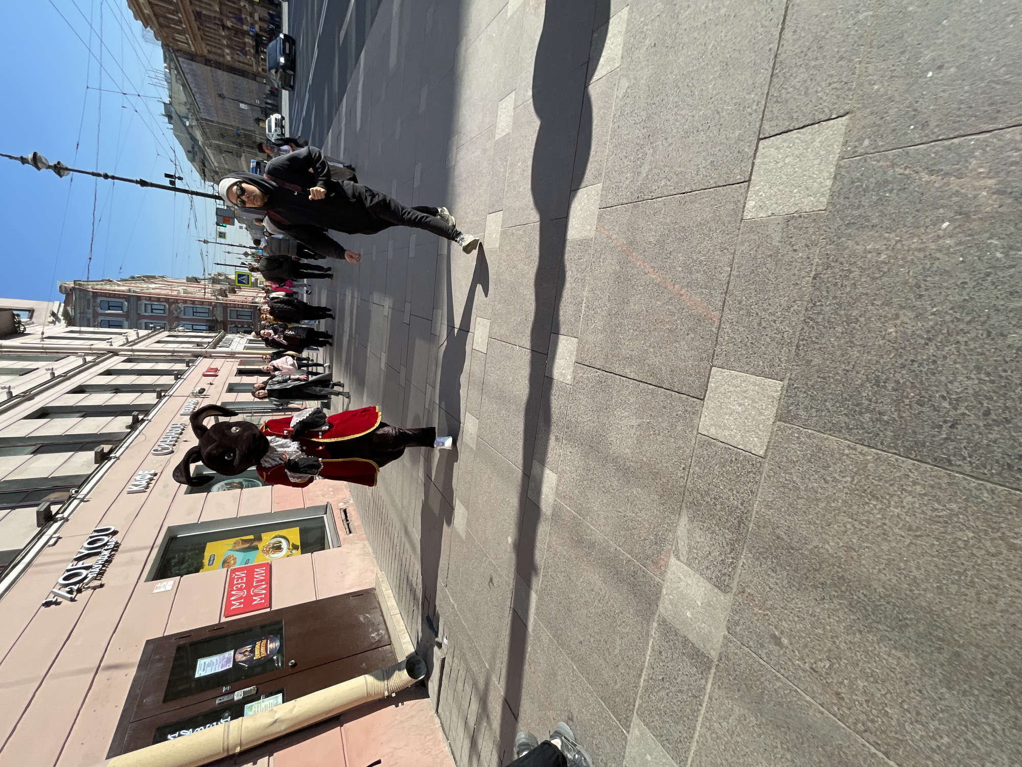 On a sunny day in Saint Petersburg, Russia, a street scene unfolds. A dark-haired man wearing sunglasses and a black hooded jacket walks at a moderate pace on the sidewalk. He wears a white head covering. Several other pedestrians are behind him. A performer dressed in a large rabbit costume and a red and black costume stands on the sidewalk, turned slightly toward the street. A building with a beige facade and signs in Russian is to the left. The pavement is made of large, gray stone tiles. A long shadow stretches across the sidewalk.