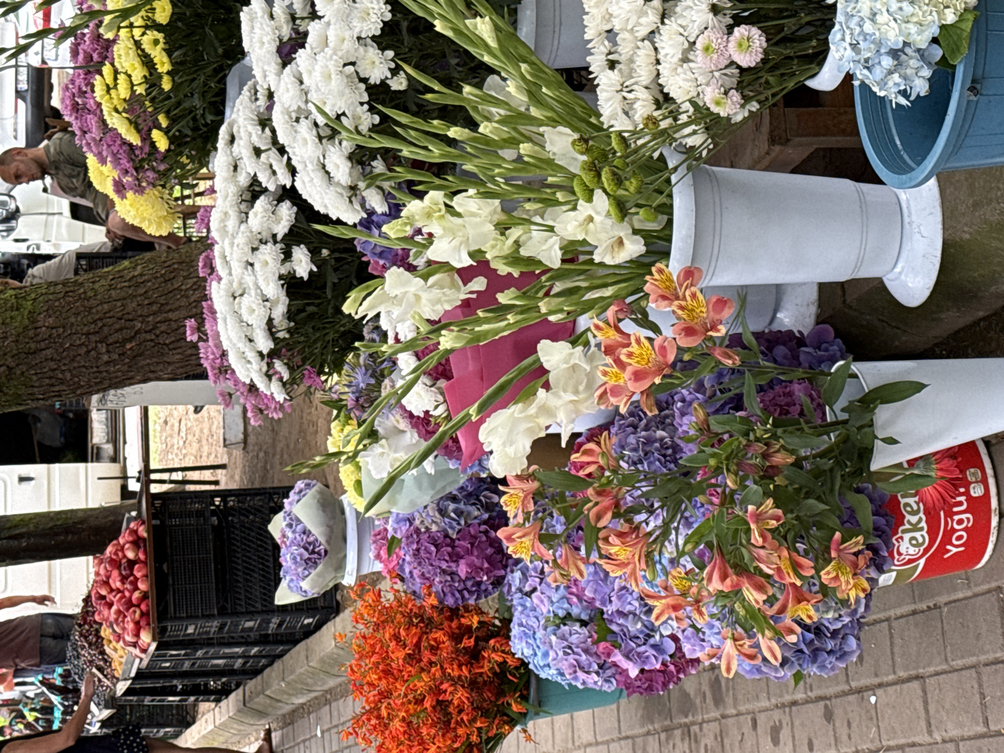 The image shows a street-level view of a flower and fruit stand. In the foreground, an array of colorful flowers are displayed in buckets and vases. These include bright orange flowers, purple and blue hydrangeas, delicate peach-colored lilies, and tall white gladiolus. Behind the flowers, more bouquets of white, yellow, and purple chrysanthemums are visible. To the left of the flowers, a fruit stand displays piles of red apples and dark berries. A person's legs are visible to the far left, wearing a dark skirt and sandals, suggesting they are a shopper. Further back, a man sits on a bench, observing the scene. The background reveals a white van and more of the market environment. The ground is paved with brick-like stones. The scene appears to be outdoors, with a tree trunk visible on the right side of the image.