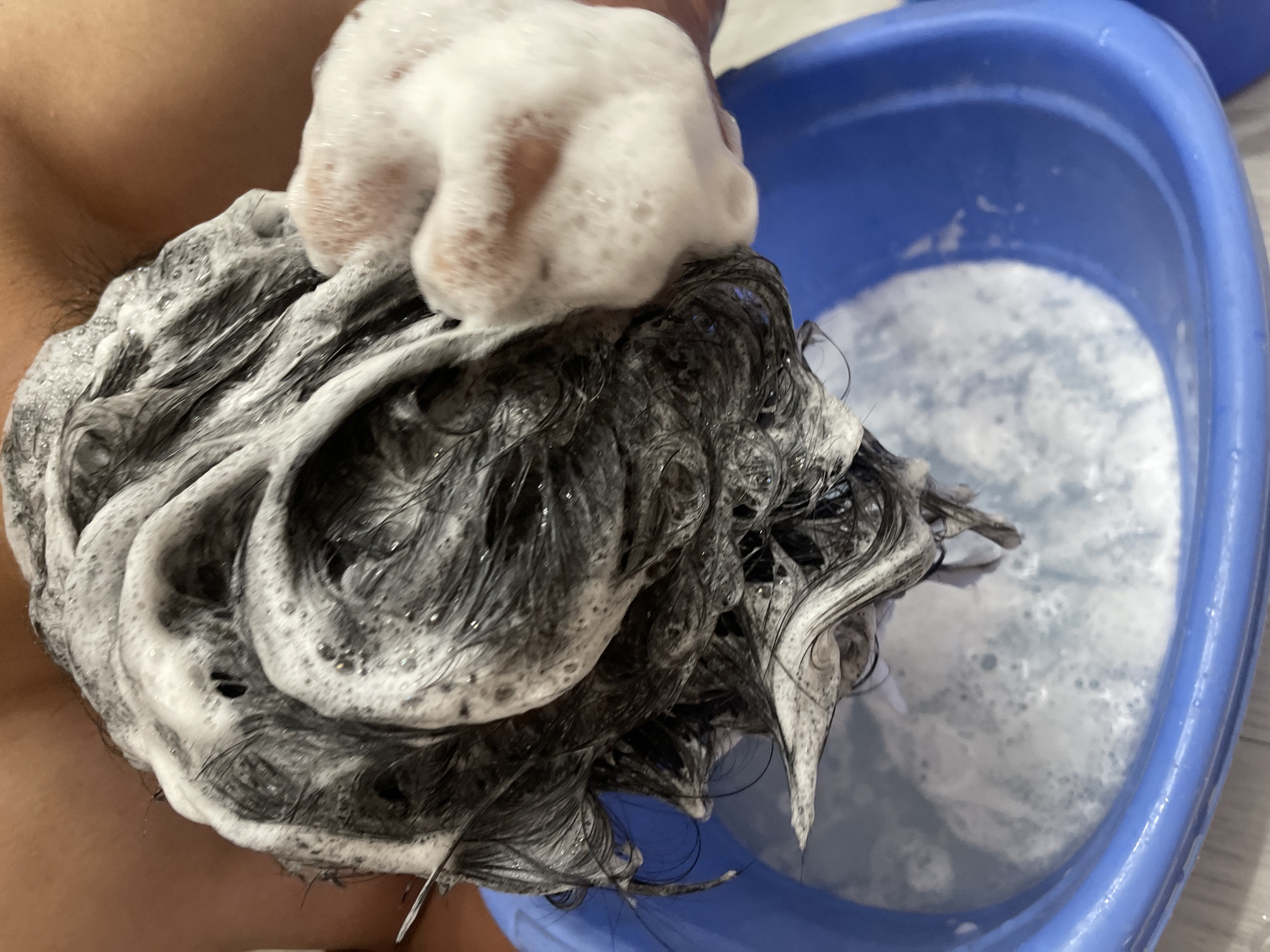 The image shows a close-up view of a person washing their hair in a blue plastic basin. The hair is dark and appears to be lathered with shampoo, creating white foam. A hand, also covered in foam, is actively massaging the scalp. The basin contains soapy water, and the background is blurry, suggesting an indoor setting. No specific location in Bishkek City, Kyrgyzstan, can be identified from the provided image.