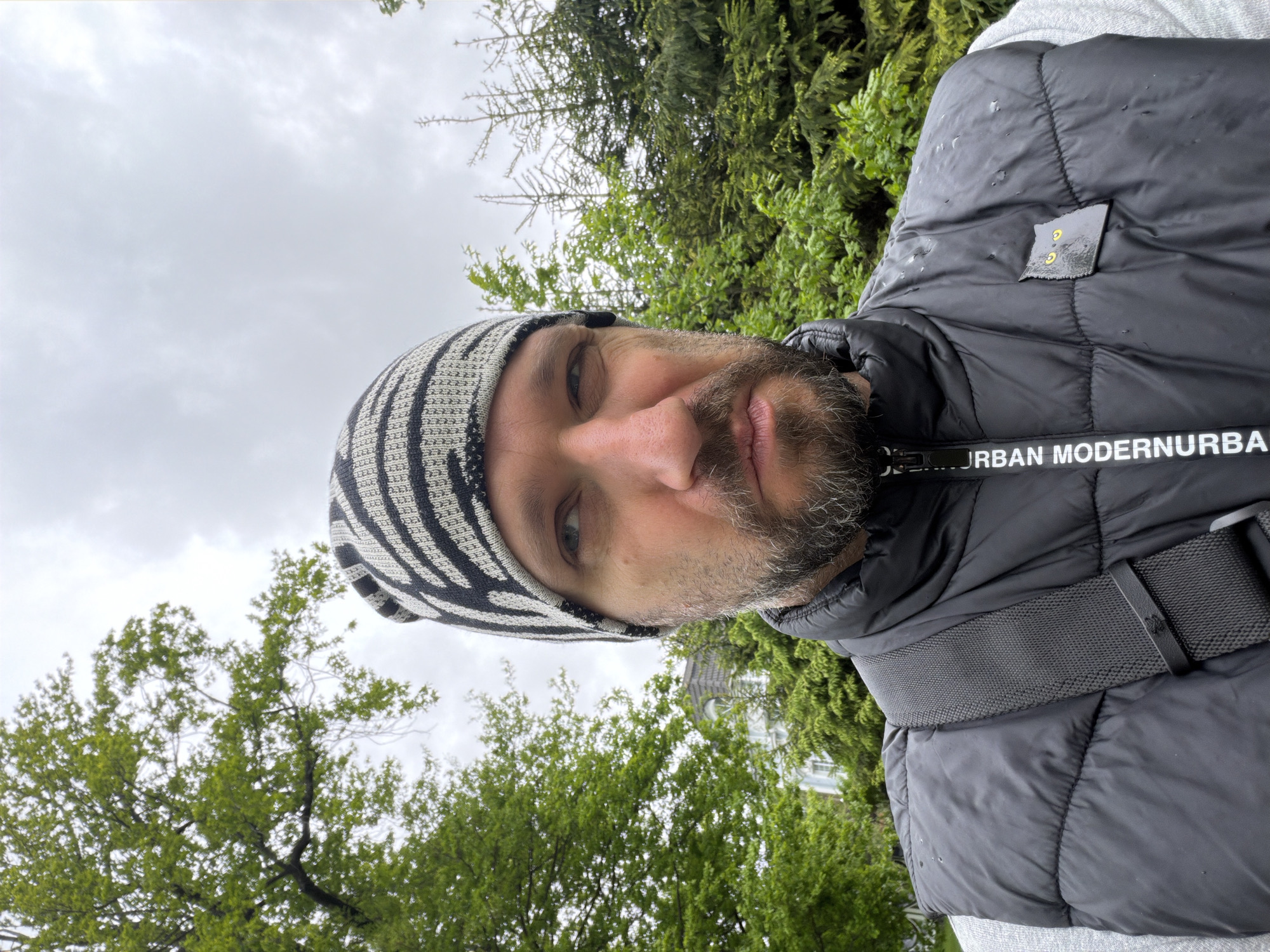 The image is a selfie of a man with a beard and a striped beanie, wearing a black puffer vest over a gray shirt. The vest has a strap across the chest and a small patch on the upper left. The man appears to be outside, with trees and a cloudy sky visible in the background. Based on the architecture visible through the trees, the location is Strelna, Russia.