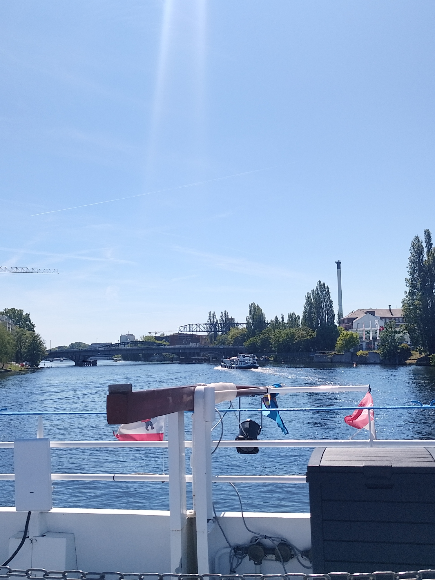 A daytime shot from a boat on a river with a blue sky overhead. There are several bridges in the background and some buildings and trees on the banks of the river. A boat is moving on the water in the middle ground. In the foreground, there is a railing of the boat, with flags and some equipment attached to it. The river seems to be in the city of Unknown.