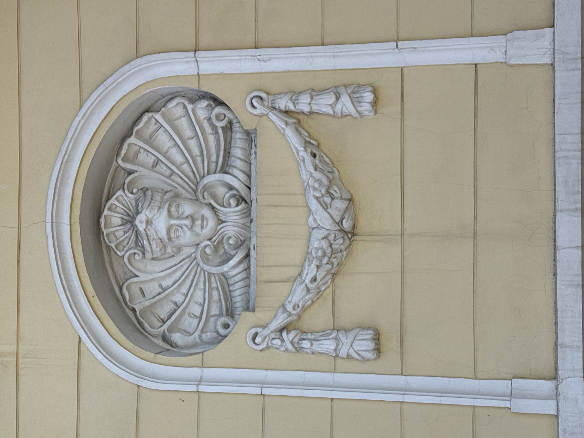 The image shows a decorative bas-relief sculpture on a yellow-painted building in Krasnoyarsk, Russia. The sculpture is comprised of a semi-circular arch containing a face surrounded by radiating shells. From either side of the face element extends a curved arm-like form terminating in a tied bundle. The sculpture is framed by a square border. Cracks are visible on the painted wall surface.