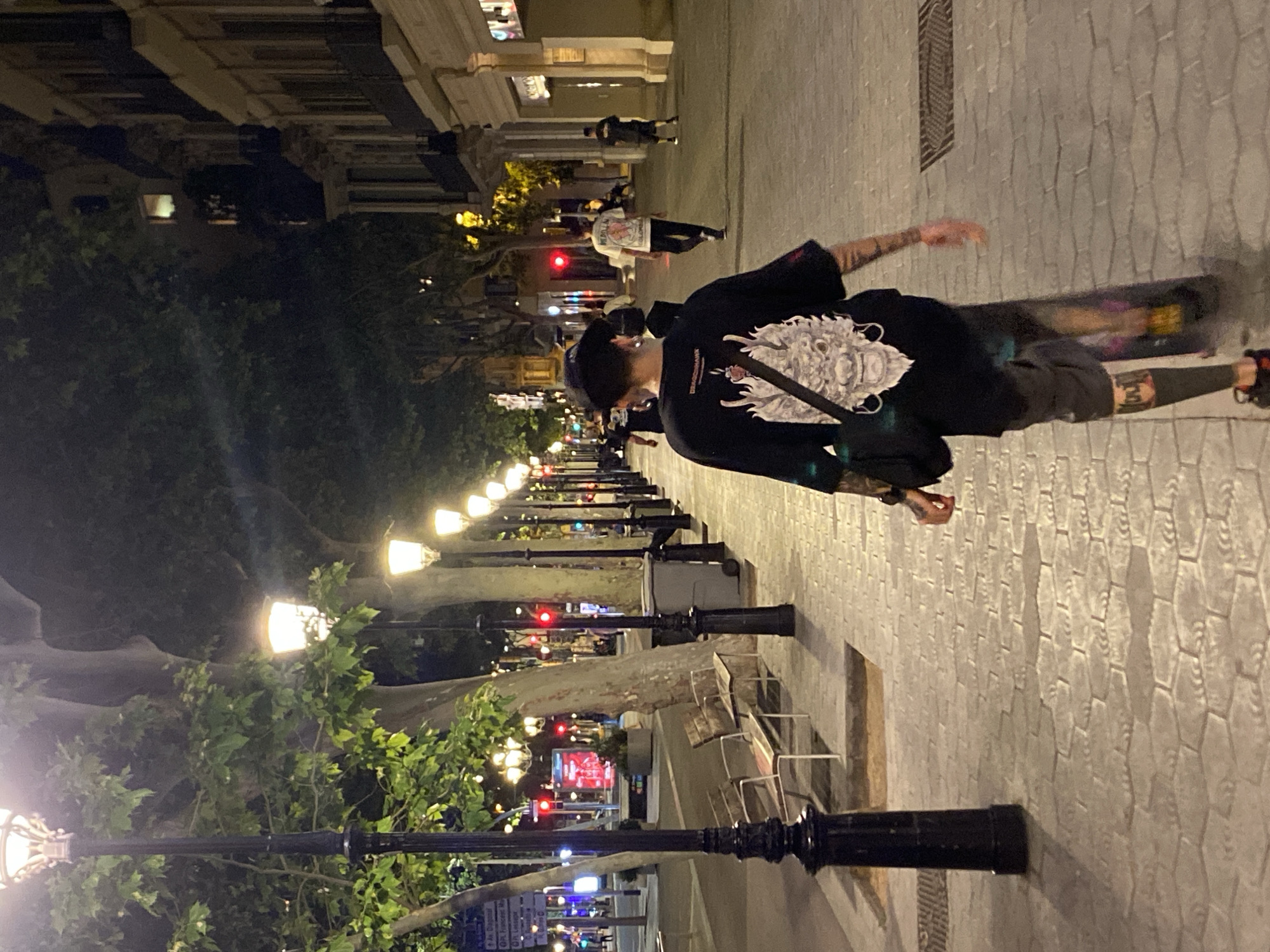 Captured at night, the scene shows a city street, possibly in Barcelona, Spain, illuminated by a series of streetlights that stretch into the distance. Trees line the street, adding to the ambiance. In the foreground, a person wearing a black jacket with a prominent white graphic on the back walks along the sidewalk. Other pedestrians are visible further down the street, suggesting a moderately busy night scene. The street is paved with a patterned surface, and architectural details of buildings can be seen in the background, adding depth to the urban environment.