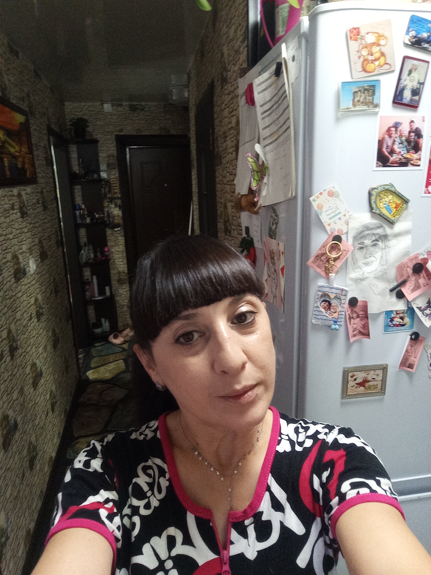 A woman with dark, short hair and bangs poses for a selfie in what appears to be a hallway in a residential apartment in Belogorsk, Russia. She is wearing a black and white patterned shirt with pink trim. To her right is a white refrigerator adorned with various magnets, photographs, and a drawing. The hallway walls are textured, resembling stone. A dark doorway is visible in the background, as well as a tall shelf unit filled with bottles and other items.
