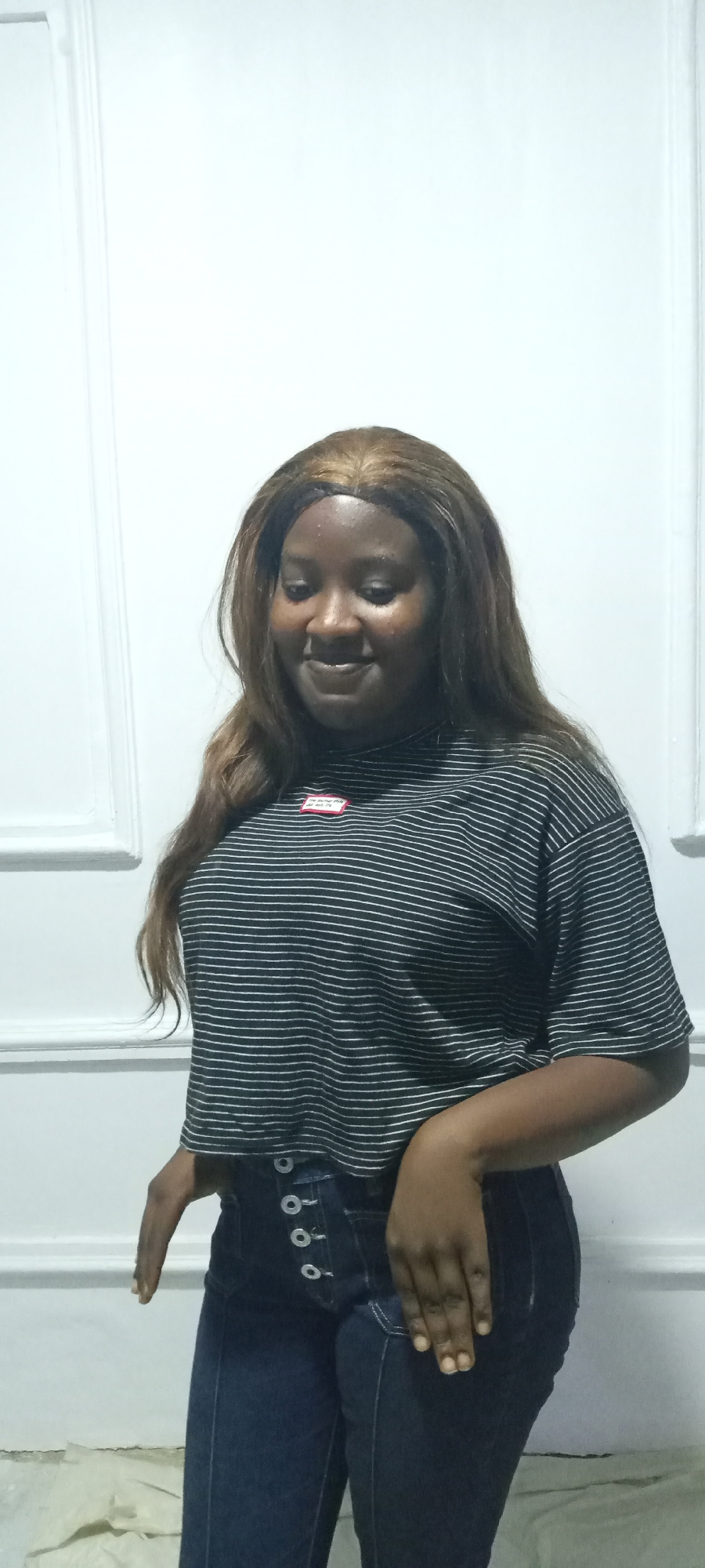 A young adult woman with dark skin and long, wavy brown hair, possibly a wig with a visible parting, is depicted in an indoor setting in Jos, Nigeria. She stands against a plain white wall that features subtle white decorative paneling or trim. Her gaze is directed downwards, and a gentle, subtle smile plays on her lips, conveying a sense of calm or slight shyness.
She is casually dressed in a black and white horizontally striped, short-sleeved crop top. A small, rectangular red label with indistinct white text is visible on the upper left side of her chest. She wears dark blue, high-waisted jeans characterized by a series of prominent silver-toned buttons running down the front fly. Her right hand is casually placed on her hip, with her fingers slightly curled.
The scene is lit by artificial light, as indicated by the absence of natural light cues and some glare on the wall and her forehead. The image captures the woman in a posed, static moment.