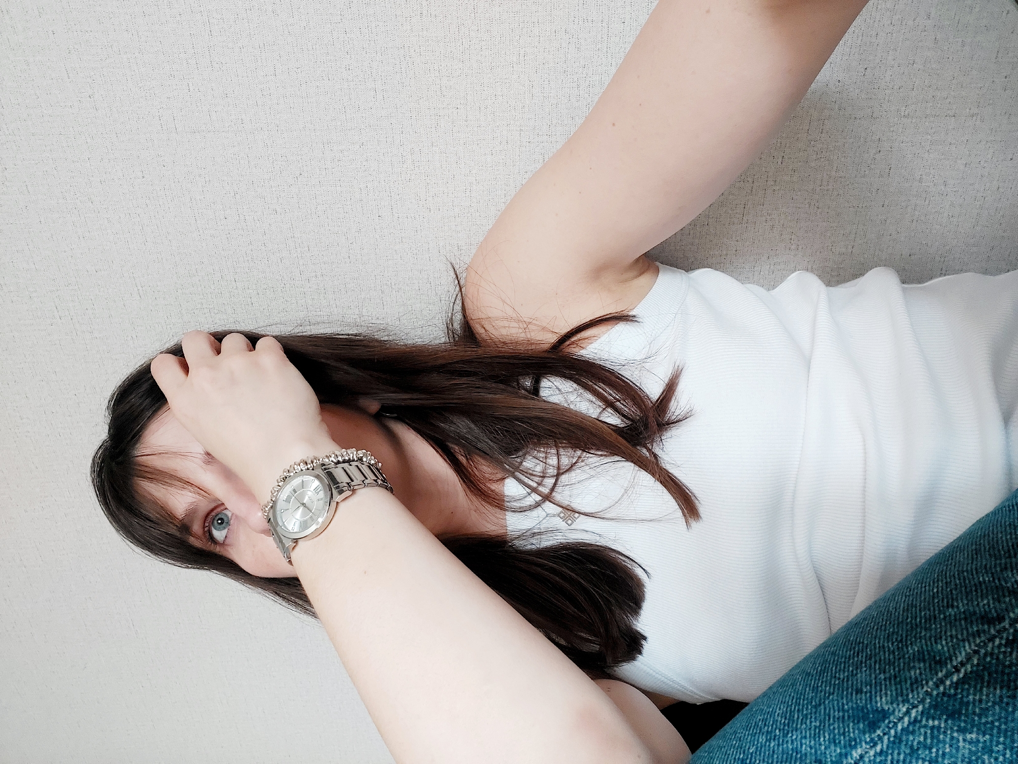 The image shows a woman indoors, likely in Chaikovsky, Russia. She is positioned diagonally across the frame, with her head angled towards the lower-left corner. She has long, dark brown hair, light skin, and blue eyes. Her right hand is raised towards her forehead, with her fingers touching her hair. A silver wristwatch with a bracelet-style band is on her right wrist. She is wearing a white ribbed tank top and blue denim jeans. The background is a textured, light-colored wall.