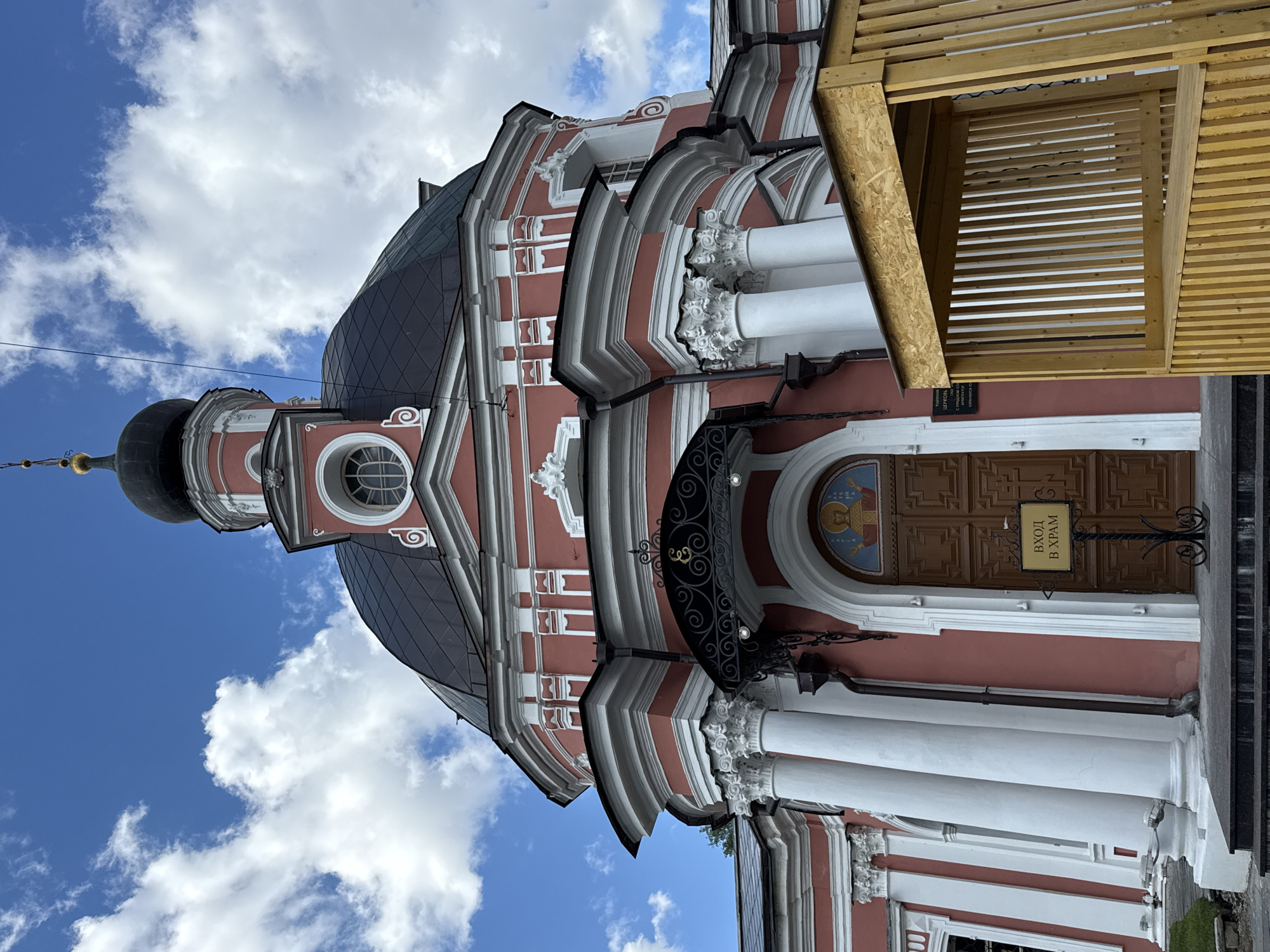 The image shows a pink and white domed church with a black dome, under a blue sky with white clouds. A wooden structure is in the foreground to the right. The entrance to the church has a wooden door and a sign that says "ВХОД В ХРАМ" (Entrance to the temple). The church is located in Moscow, Russia.