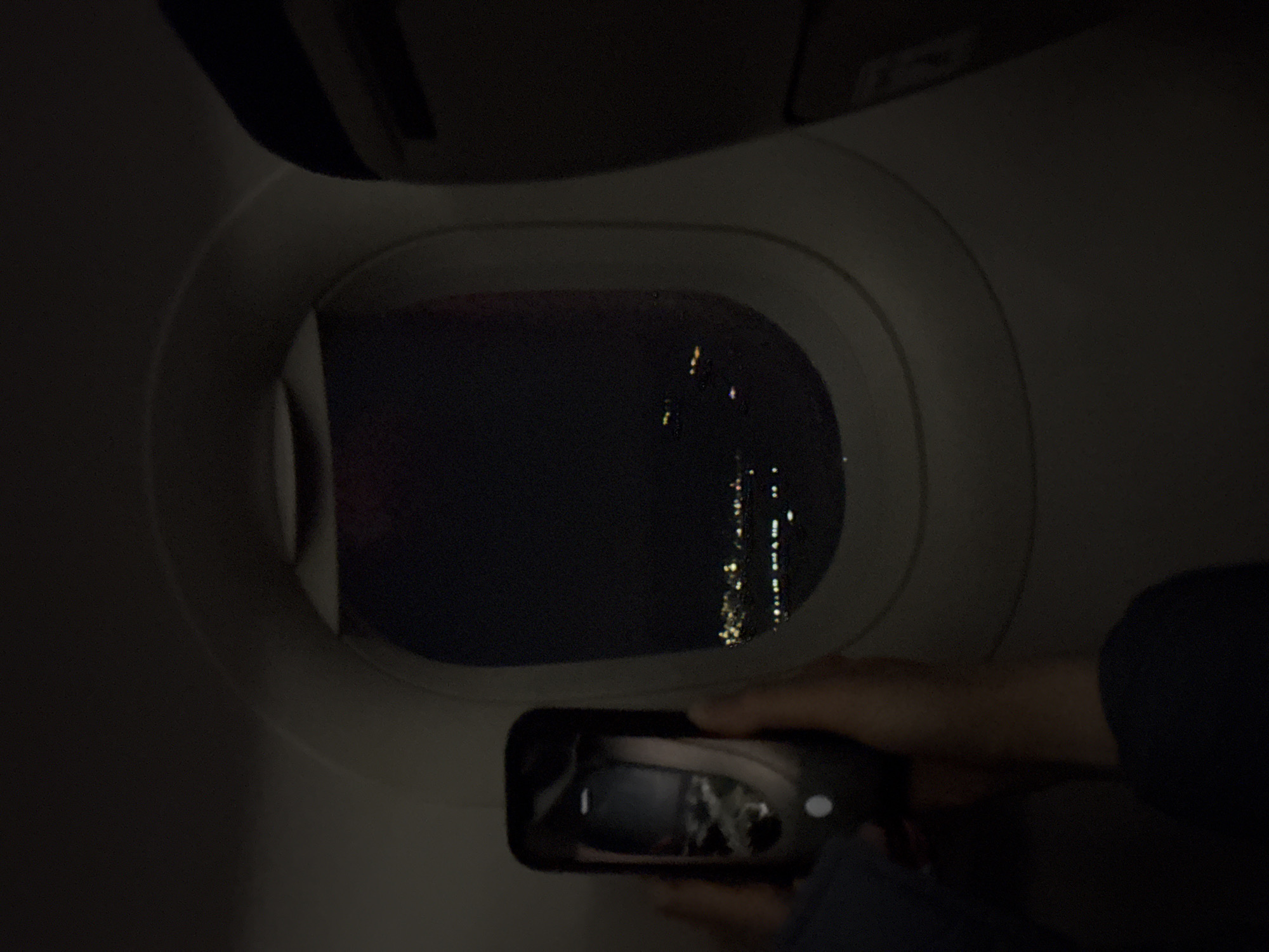 An interior view from an airplane at night shows a person holding a smartphone, aiming it towards the oval-shaped window. The person's hands are visible at the bottom right of the frame, gripping the phone. Through the airplane window, a dark night sky is visible, interspersed with numerous, streaky city lights appearing as vertical lines of yellow and white dots in the distance. The smartphone's screen displays a reflection, possibly of the cabin interior or the window itself, indicating the person is likely capturing the night view. The dim lighting within the cabin illuminates parts of the window frame and an overhead panel. The scene captures the activity of documenting the night sky and urban sprawl from a flight over what is contextually identified as Mayisyan, Armenia. The time of day is clearly night, with no specific weather conditions or visible text discernible in the image.