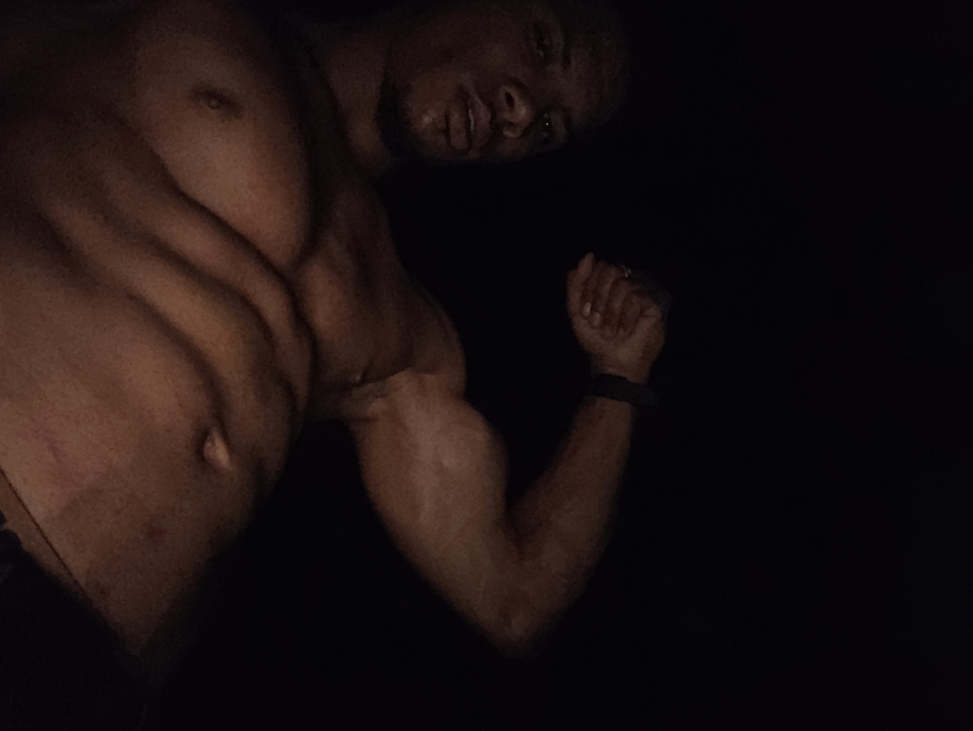 The image shows a close-up, low-angle shot of a muscular individual with dark skin and short dark hair, viewed from the chest up. The subject is leaning forward, looking down and to the left, with their mouth slightly open. Their left arm is flexed and raised, with a black band visible on their wrist. The background is completely black, obscuring any environmental details. The lighting appears to be dim, highlighting the musculature of the subject's chest and arm. There are no discernible landmarks or features that would indicate a specific location within Varna, Bulgaria, or any other city.