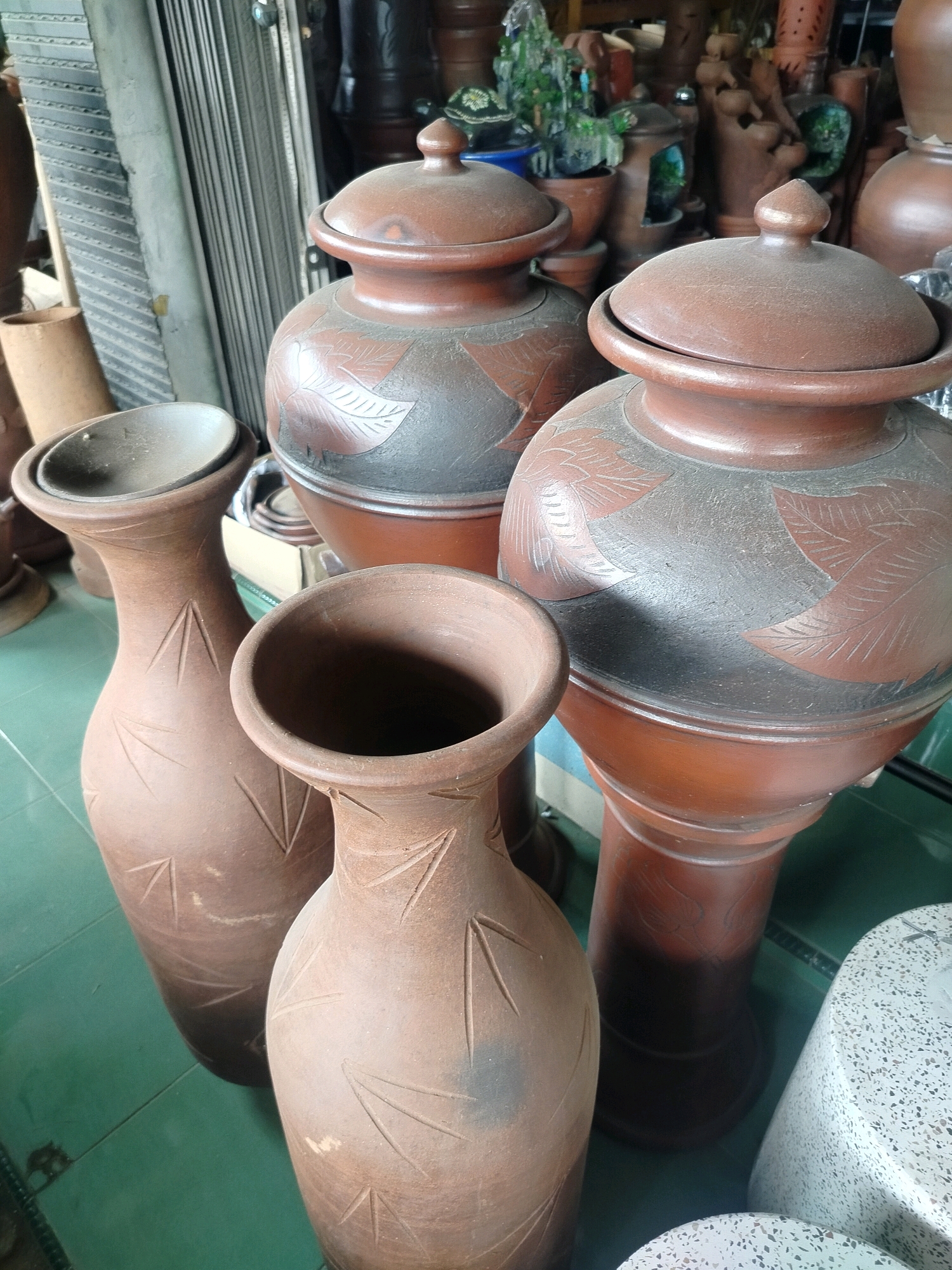 The image displays a collection of large ceramic pots and vases, predominantly in shades of brown and terracotta, displayed outdoors on a tiled green floor. In the foreground, three distinct vases are visible. The largest, on the right, has a wide opening and a textured surface with etched linear patterns. To its left is another similarly shaped vase, slightly smaller, also with etched patterns. To the far left, a third, taller vase with a narrower neck and wider base is present, exhibiting similar etched designs. In the background, several larger, lidded pots are featured. One prominent pot on the right is decorated with leaf-like patterns etched into a darker, matte surface. Another similar pot stands to its left. Behind these, a variety of other ceramic items, including smaller pots and possibly figurines, are arranged in what appears to be a market stall or shop. The environment suggests an outdoor commercial setting, possibly in the city of Kroya, Indonesia, given the prevalence of such crafts in the region. The lighting indicates daytime, with some shadows suggesting overhead cover or placement. No people are visible in the frame.
