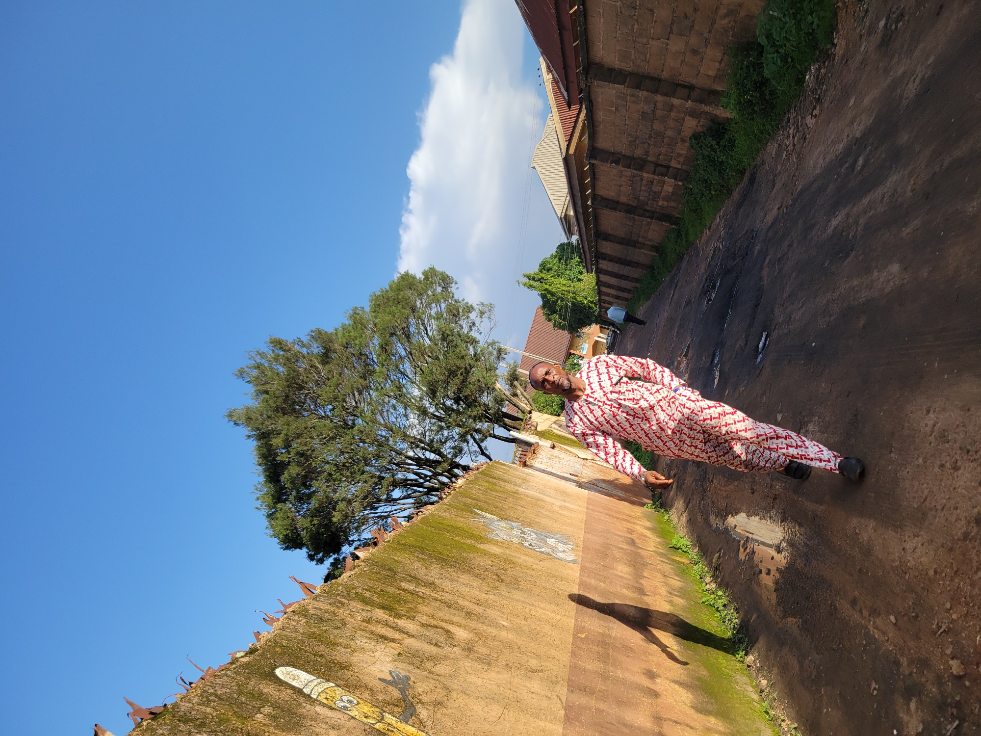 In a sunlit scene in Jos, Nigeria, a man in a red and white patterned traditional outfit walks down a dirt road lined by brick walls and a moss-covered concrete wall. The concrete wall on the left features graffiti, including a cartoonish drawing of a pencil. The man casts a long shadow on the wall, indicating the low angle of the sun. Behind him, other buildings and trees are visible. The sky is a vibrant blue with scattered white clouds.