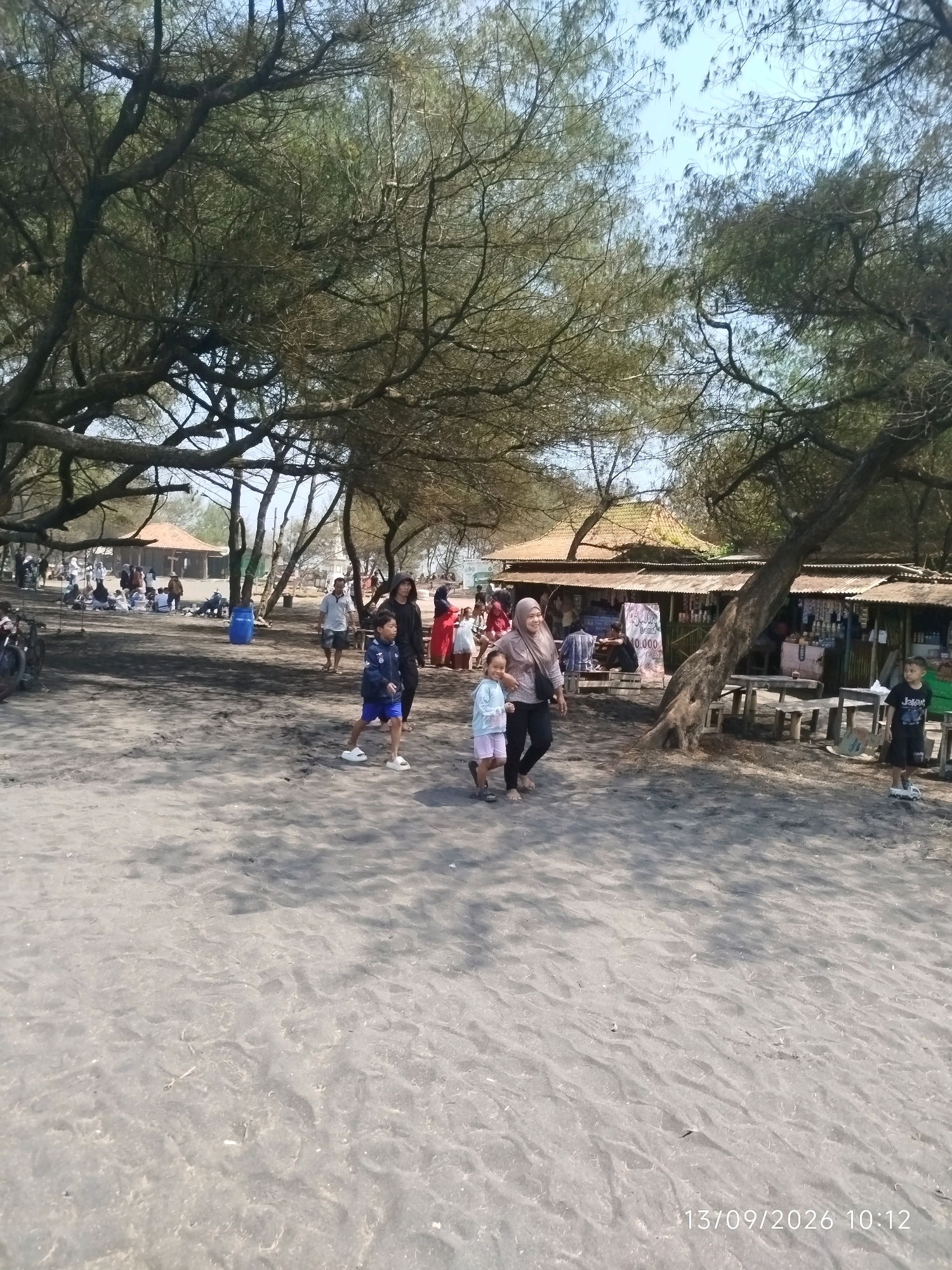 In Pleret, Indonesia, a group of people are gathered on a sandy beach shaded by trees. The sand appears dark, possibly volcanic or mixed with other dark particles. In the foreground, a young boy in a blue and white jacket and blue shorts walks towards the left, wearing white slides. Behind him, a woman in a light-colored shirt and dark pants, wearing a headscarf, walks with a young girl in a light-colored shirt and pink shorts. To the right, another young boy in a black t-shirt and shorts stands on the sand, with a toy vehicle at his feet. In the middle ground, several people are seen walking and sitting, with stalls or small shops visible behind them. These structures have roofs and displays of goods. Further in the background, more people are visible, along with some tents or temporary shelters and bicycles. The trees overhead have a sparse canopy, allowing sunlight to filter through. The sky is clear and blue. A date and time stamp "13/09/2026 10:12" is visible in the lower right corner of the image.