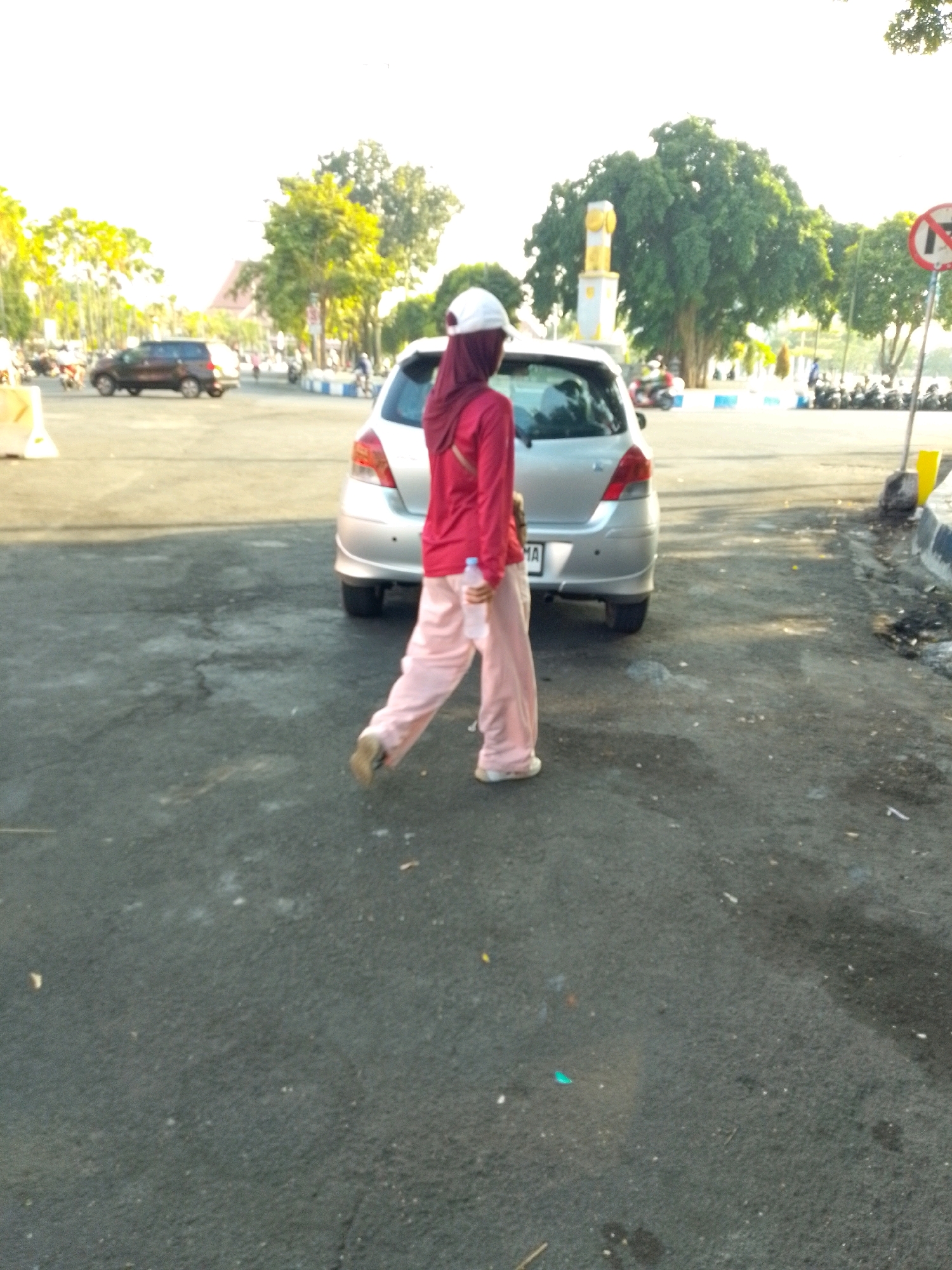 A woman wearing a pink hijab and light pink pants walks away from the camera, holding a water bottle in her right hand. She is walking on a paved road, with a silver car parked to her left. In the background, there are trees, a monument, and parked motorcycles. The scene appears to be outdoors in a public area, possibly a park or a street, with buildings visible in the distance. The lighting suggests it is daytime, likely late afternoon or early morning, given the long shadows. This location is identified as being in the city of Jember, Indonesia, specifically near the Alun-Alun Jember, a central town square known for its iconic landmark, the Tugu Lampu Al-Alun.