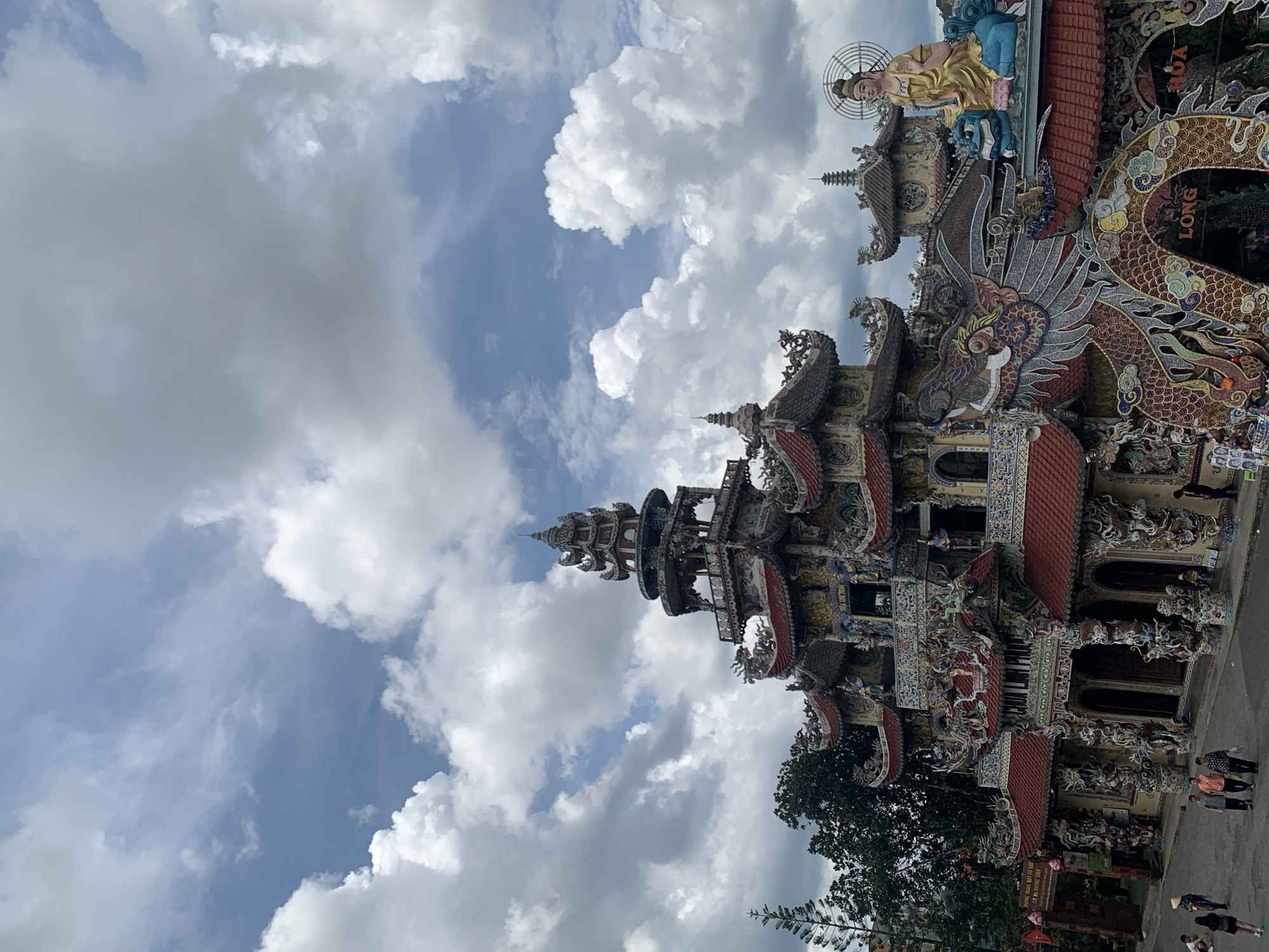 An exterior, eye-level, medium shot captures the colorful and ornate architecture of the Da Lat Crazy House in Xuân Thành, Vietnam, under a bright, cloudy sky. The building, a fantastical, multi-tiered structure resembling a whimsical castle, is adorned with intricate ceramic mosaics depicting dragons, mythological creatures, and floral motifs. Red-tiled roofs crown various sections of the building, which features numerous balconies, windows, and spires. To the right of the building, a large, golden statue of Buddha sits atop a blue creature, radiating a serene presence. The foreground shows a paved walkway with several individuals strolling and observing the structure. Trees and lush greenery frame the sides of the building, indicating a natural setting. The overall scene is vibrant and detailed, highlighting the unique architectural style of the Crazy House.