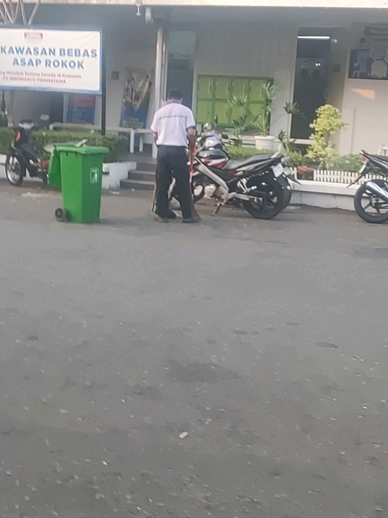 In Trihanggo, Indonesia, a person is standing near a green trash bin and a motorcycle. The person is wearing a white shirt with a collar and dark pants. They appear to be in the process of urinating into the trash bin. The background shows a building with several windows, plants, and a white picket fence. The ground is paved with asphalt, and there are other motorcycles parked in the vicinity. A banner on the building reads "KAWASAN BEBAS ASAP ROKOK," which translates to "SMOKING-FREE AREA." The overall scene suggests a public or semi-public space where the individual is disregarding the posted smoking ban and public decency.