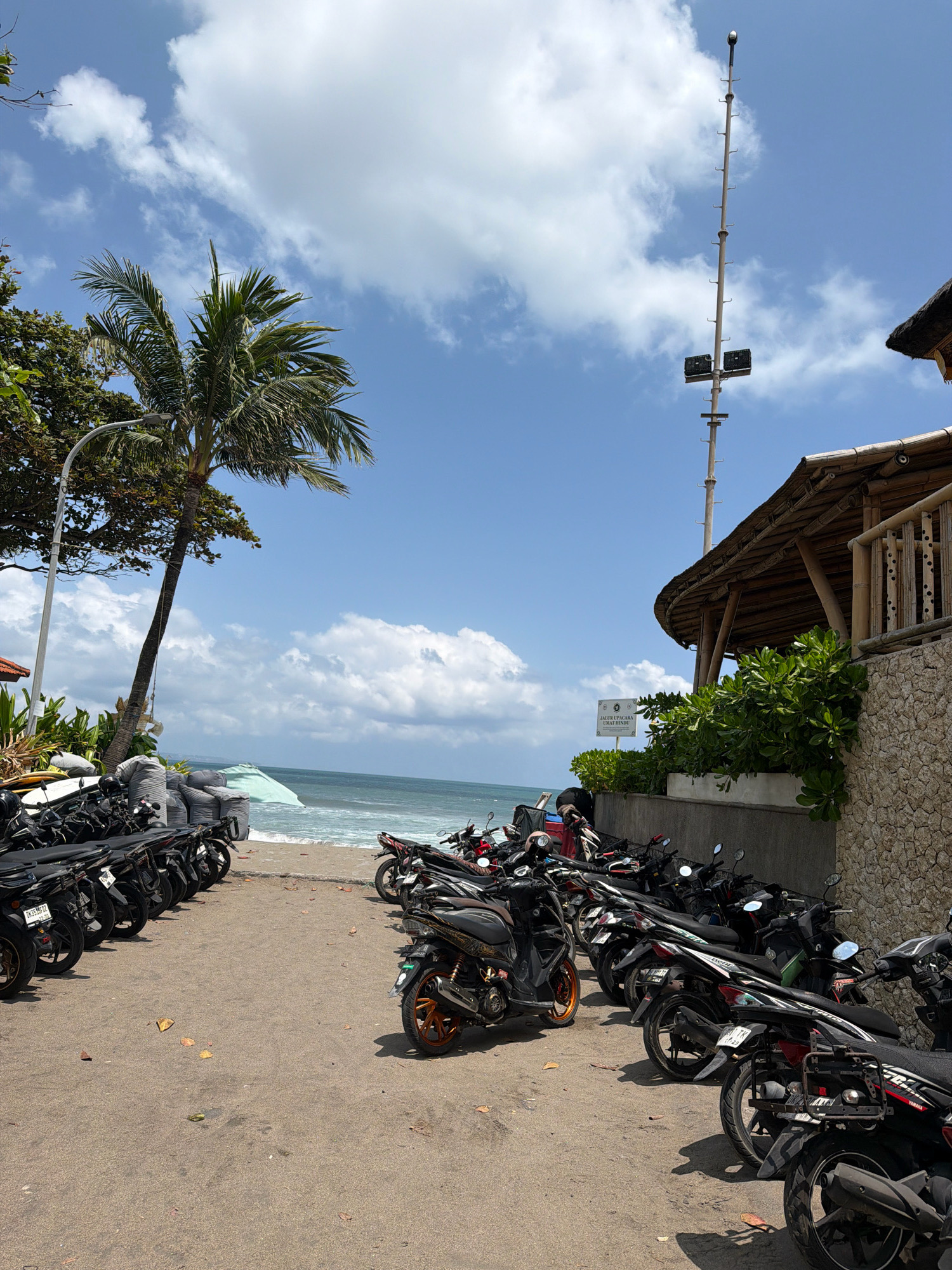 The image captures a street scene in North Kuta, Indonesia, near a beach. In the foreground, a sandy parking area is filled with numerous parked motorcycles of various colors, predominantly black. Some motorcycles have visible license plates. Behind the motorcycles, a beach stretches out with gentle waves meeting the shore. Further back, a body of water is visible, with a hazy horizon. To the right, a building with a thatched roof and bamboo accents is situated, its stone wall and lush green plants adding to the tropical ambiance. A tall pole with lights and antennas stands against a bright blue sky with scattered white clouds. A palm tree leans towards the left side of the frame, near a street lamp. The overall scene depicts a public space likely frequented by beachgoers and locals.