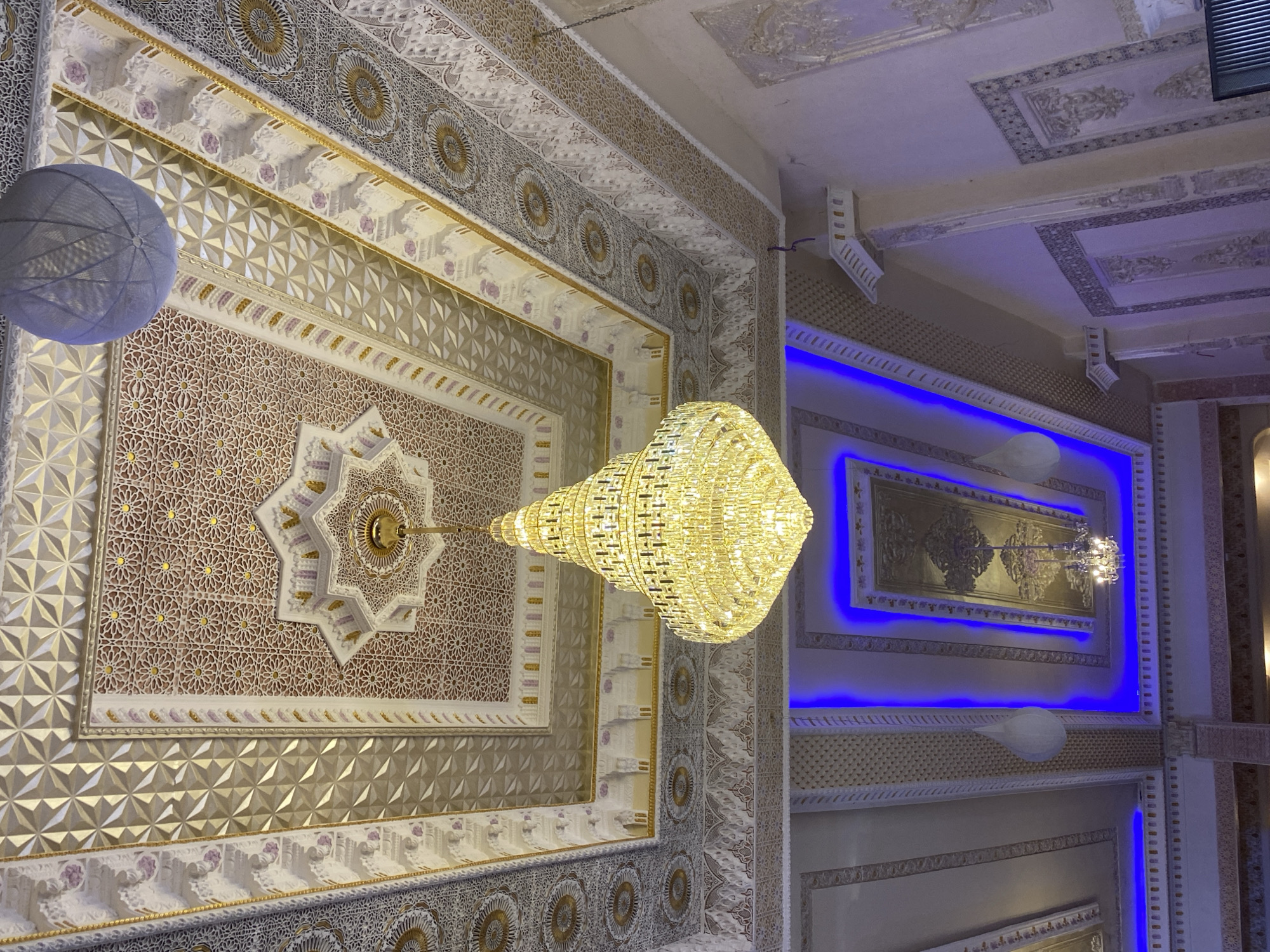 The image depicts the interior ceiling of a building, likely a mosque or a grand hall, in Barakin Lerawi, Nigeria. The ceiling is elaborately decorated with intricate geometric patterns and ornate carvings, featuring a dominant star motif in the center. A large, multi-tiered crystal chandelier hangs prominently from the ceiling, casting a warm, golden light. In the background, recessed lighting with a vibrant blue glow illuminates further sections of the ceiling, highlighting more decorative panels and a smaller chandelier. There are also decorative spherical light fixtures hanging from the ceiling. The walls appear to be adorned with decorative panels and pilasters, suggesting a richly ornamented architectural style. No people are visible in the image. The scene is static and focuses entirely on the architectural and decorative elements of the space.