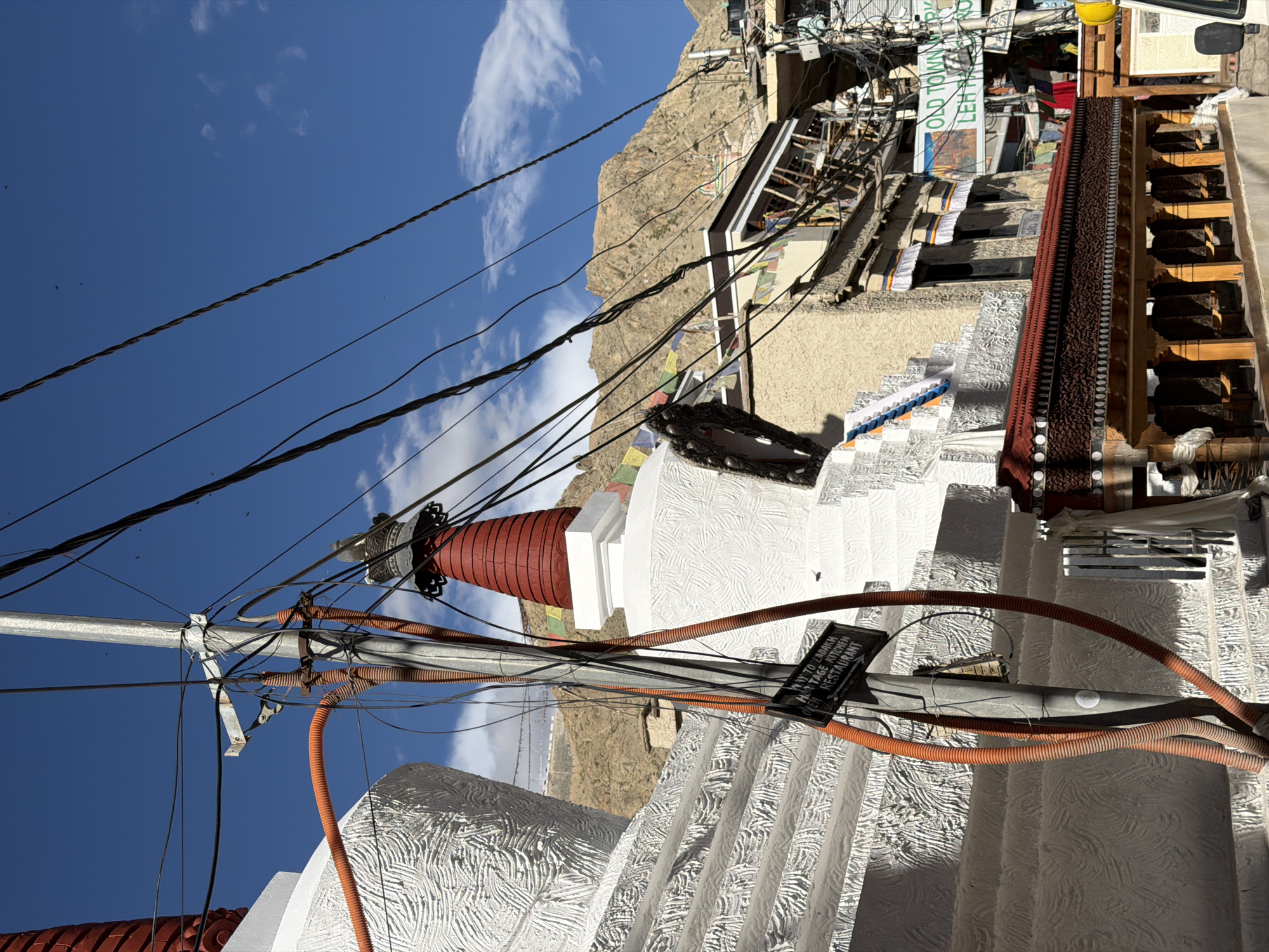 In the city of Leh, India, on a sunny day with a blue sky and scattered clouds, a street scene unfolds. A tall, gray metal pole, laden with a dense network of black electrical wires, stands prominently in the foreground. Two ribbed orange conduits run vertically along the pole. To the left, a large, white stupa with a textured surface and a red conical top is partially visible. To the right, another white stupa, also with a red conical top, is more fully in view. Below the stupas, a structure with intricate wooden carvings and a red tiled roof is visible, with prayer wheels lining its base. In the background, buildings with a light beige facade are clustered together, adorned with prayer flags. A sign reads "OLD TOWN PARK LEH." The overall impression is one of a busy urban environment where traditional structures coexist with modern infrastructure, such as the abundant electrical wiring.