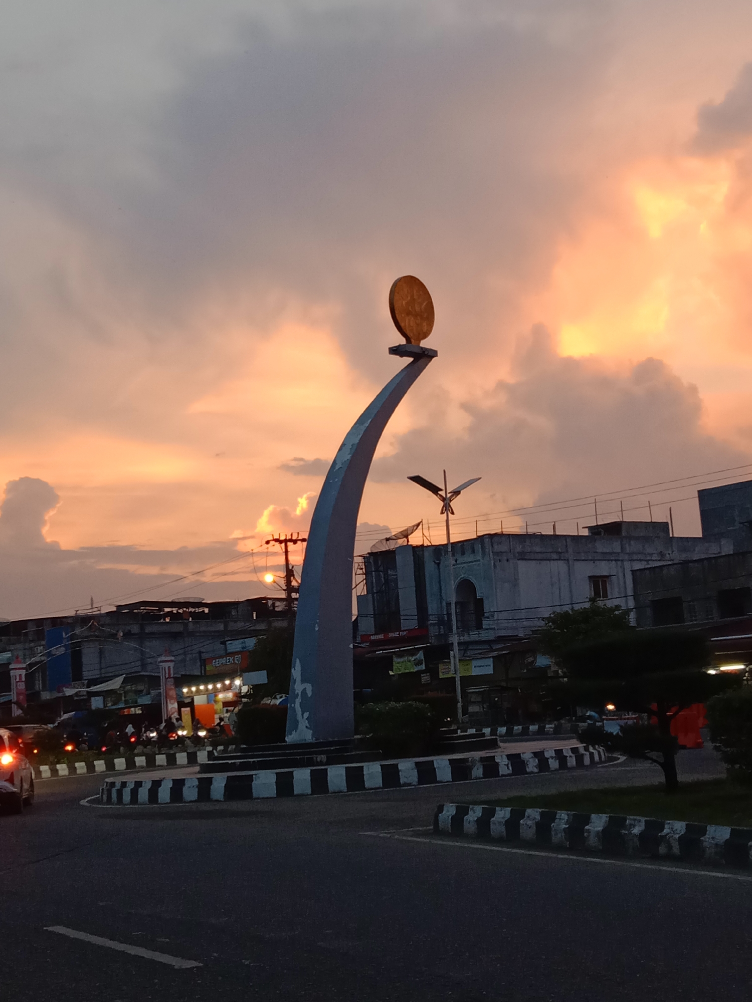 This full-shot photograph captures a city roundabout in Lhokseumawe, Indonesia, during the golden hour, likely sunset, characterized by a dramatic sky filled with warm orange, pink, and gray clouds. A prominent monument dominates the center of the roundabout, featuring a large, golden, circular emblem with an intricate pattern, set atop a gracefully curved gray and blue pedestal.
The road surrounding the monument is clearly defined by black and white striped curbs. Vehicles, including cars and motorcycles with their lights on, indicate active traffic, with a few people visible on motorcycles. Low-rise buildings, appearing to be commercial or residential establishments, line the streets, with power lines visible crisscrossing above them. Artificial lights from these establishments and streetlights begin to illuminate the scene as daylight fades. A sign on one building clearly displays the word "GEPREK." The overall scene blends urban activity with the natural beauty of the evening sky.
