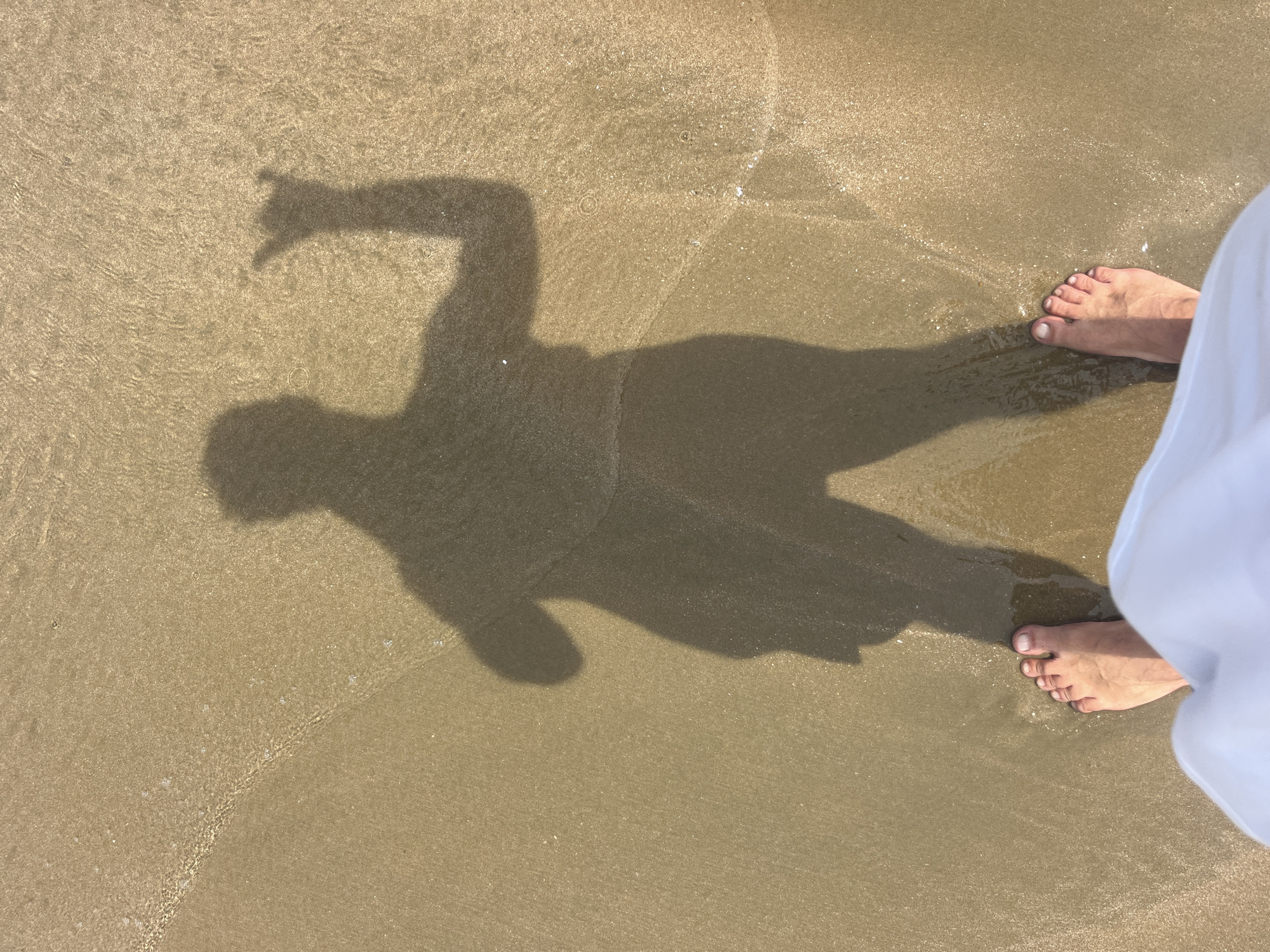 A person's shadow is cast on wet sand near the shoreline, depicting them making the "shaka" hand gesture. The person's bare feet, partially submerged in shallow water, are visible in the foreground, along with the edge of white fabric, likely clothing. The scene takes place on a beach in Mui Ne, Vietnam, characterized by its wet sand and gentle waves.