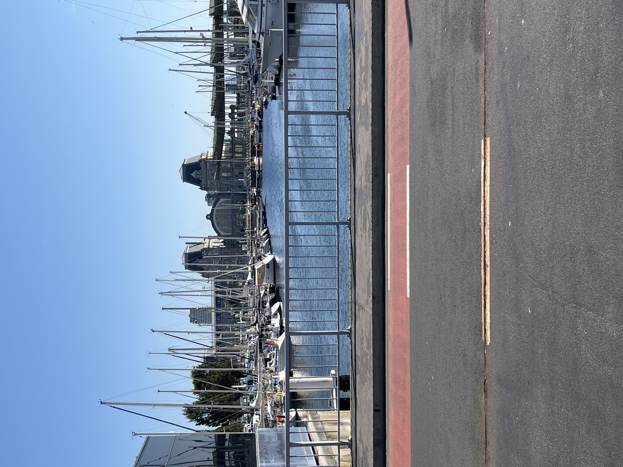 The image shows a sunny day in the port of Ostend, Belgium. In the foreground, there is a paved road with a red lane marker and a white line. Beyond the road, there is a metal railing, and behind that, a body of water filled with numerous sailboats. Several buildings are visible in the background, including what appears to be a train station or large public building with a distinctive architectural style. The sky is clear blue.