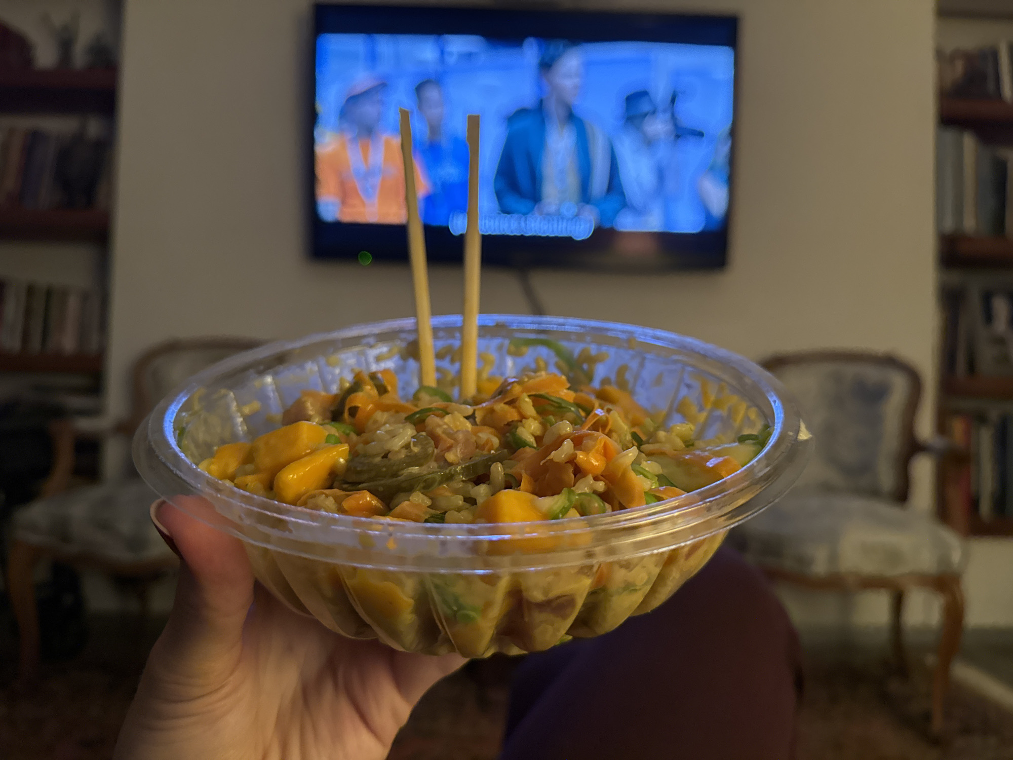 A person holds a plastic container of food with chopsticks in it in front of a television and bookshelves. The food appears to be rice-based, with sliced mango and green vegetables. The television displays a group of people. The background includes two ornate chairs and a bookshelf. The location is indeterminate and there is no indication of Mexico City, Mexico.