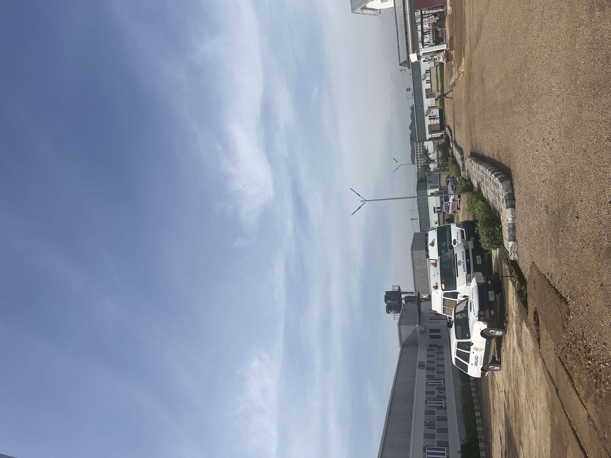 The image depicts the exterior of a medical facility, likely a hospital or clinic, in Jos, Nigeria. The scene is dominated by a large, modern building with multiple windows and an air conditioning unit visible on the exterior wall. Several ambulances, including a distinctive white station wagon with "AMBULANCE" written on its side, are parked in the lot. A man is walking across the gravel-covered area in the foreground. The sky above is partly cloudy, with a bright blue expanse and wispy white clouds. The environment suggests an active medical setting, with the vehicles indicating emergency services.