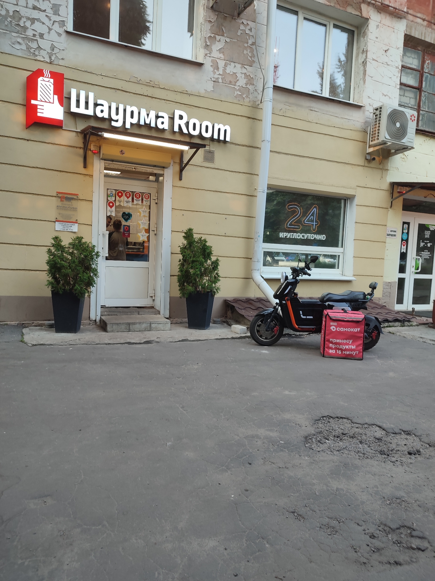 In Voronezh, Russia, on a building with a light-colored, peeling facade, a sign reads "Шаурма Room" in large silver letters. To the left of the sign is a red illuminated logo depicting a hand holding a wrapped food item. The entrance to the establishment is visible, with a person partially visible inside. Two large black planters containing green shrubs flank the entrance. To the right of the entrance, a black electric scooter is parked. A red delivery bag with the "Самокат" logo and text in Russian is placed in front of the scooter. A window displays "24" in neon orange letters, indicating 24-hour operation. An air conditioning unit is mounted on the wall above the scooter. The foreground shows a cracked asphalt ground.