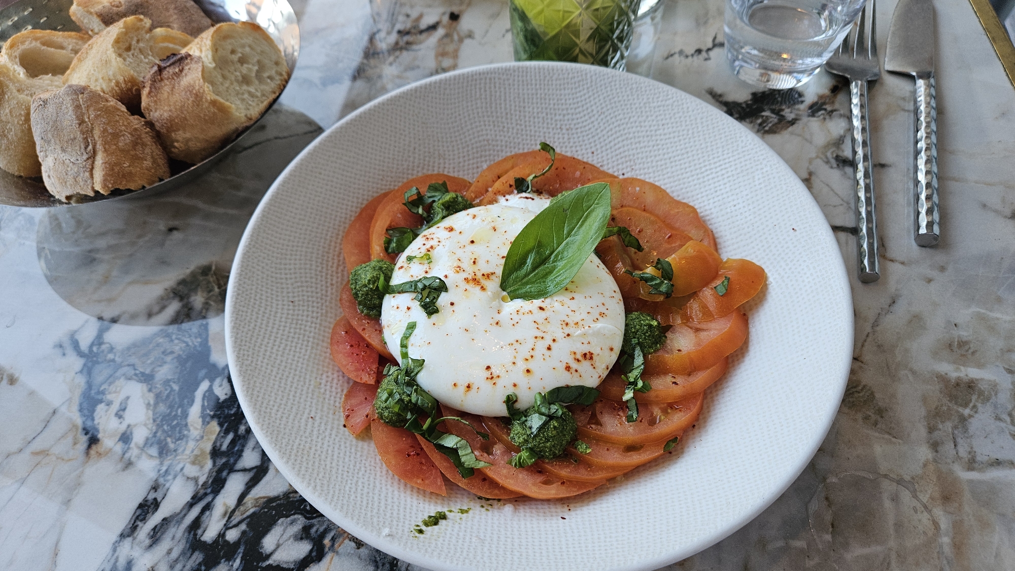 In L'Île-Rousse, France, a plate of Caprese salad is presented on a textured white ceramic dish, arranged on a marble-patterned table. The salad features thinly sliced tomatoes, a generous ball of burrata cheese topped with a basil leaf and sprinkled with paprika, and small dollops of green pesto. To the left of the plate, a silver bowl holds several pieces of bread. To the right, a glass of water and silverware, consisting of a fork and a knife with hammered handles, are placed on the table. The scene is captured from a top-down perspective, highlighting the appetizing presentation of the food and its accompaniments.