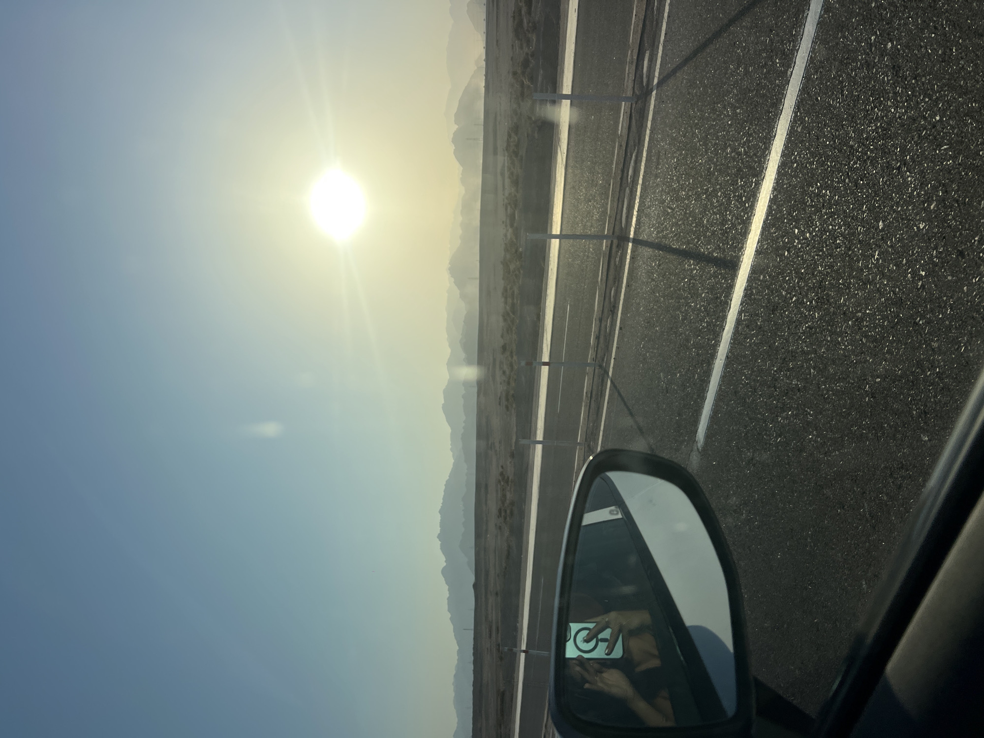A photograph taken from inside a car shows a desert landscape under a bright, hazy sun. The sun, a white orb with a halo of light, is positioned in the upper right quadrant of the sky, casting a soft glow over the scene. In the foreground, the side mirror of the car reflects a person holding a smartphone, taking a selfie. The mirror also shows a glimpse of the car's interior and the road ahead. The road itself is asphalt, with a white dividing line. Beyond the road, a sparse desert terrain stretches out, dotted with low-lying vegetation. In the distance, a range of mountains, shrouded in a light haze, forms the horizon. The sky is a pale blue, transitioning to a lighter hue near the horizon due to the sun's glare. The image depicts a typical roadside view in a desert environment, likely near Sharm El Sheikh, Egypt, given the arid landscape and mountainous backdrop often associated with the Sinai Peninsula.