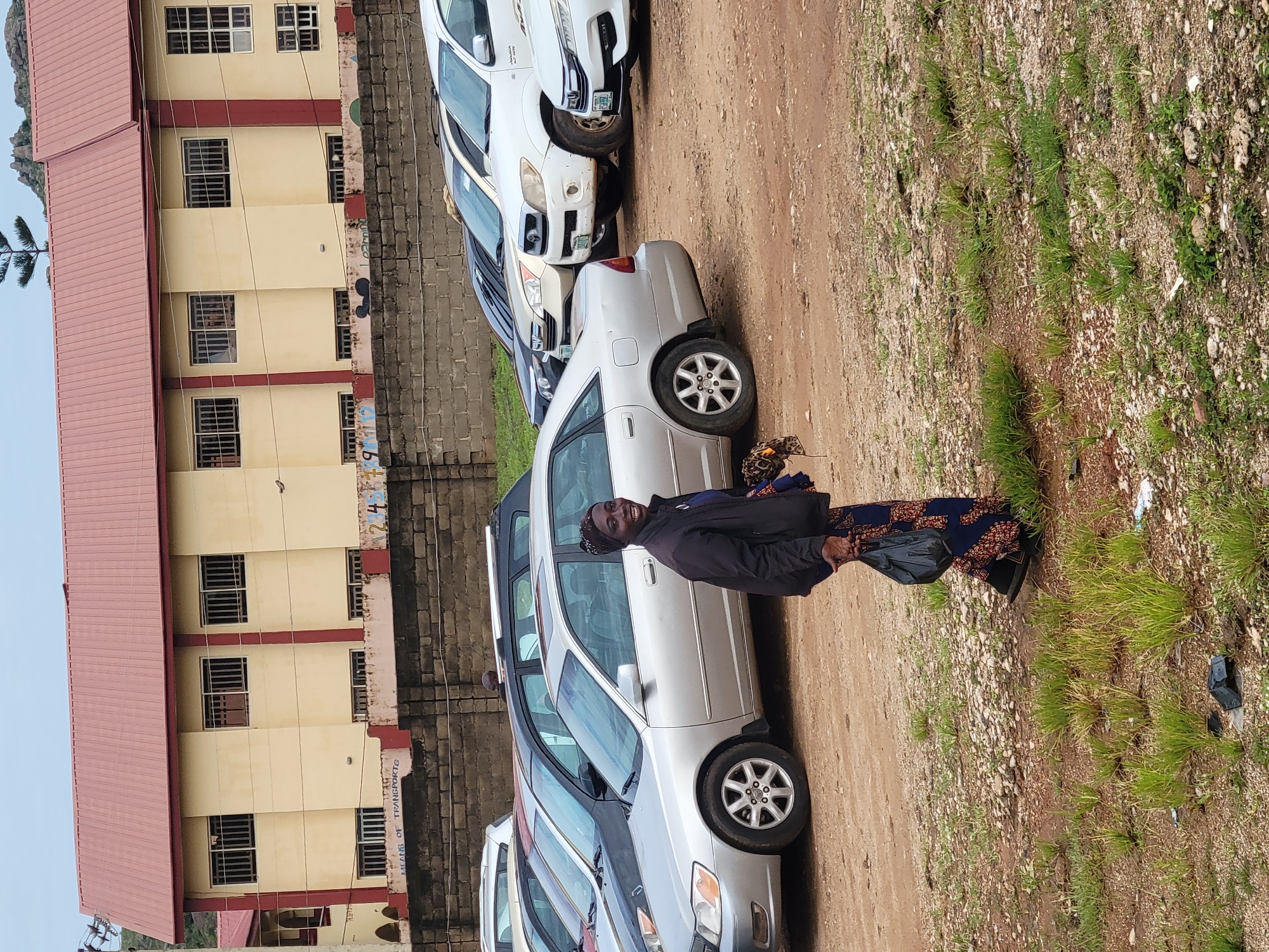 A woman wearing a black jacket over a blue and red patterned dress walks across a gravelly, outdoor area in front of a building. She has on a headscarf and is smiling at the camera. She holds a black plastic bag in her right hand and a bundle of cloth in her left. Several cars are parked behind her. The building appears to be a school with multiple windows and a red roof. The scene is set in Gura Top, Nigeria.