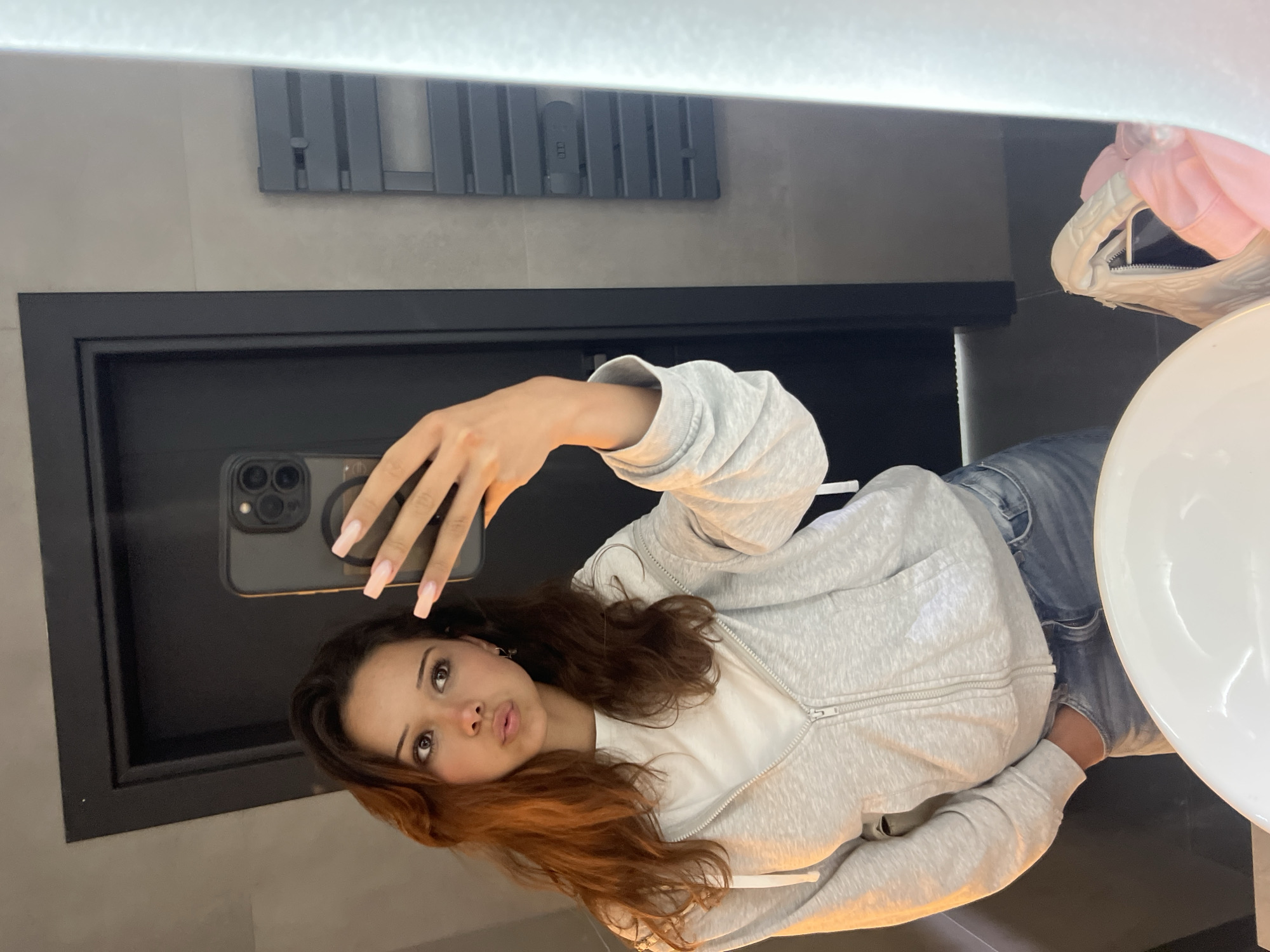 A young woman with long, brown, wavy hair takes a mirror selfie in a bathroom. She is wearing a light gray zip-up hoodie over a white t-shirt and blue jeans. Her left hand is in her pocket, and her right hand holds a smartphone, angled to capture her reflection. She has a pouty expression and is wearing long, light pink fingernails. The bathroom appears modern, with gray tiled walls, a black door, and a white sink visible in the foreground. A dark gray towel warmer is mounted on the wall to the right. The lighting is warm and slightly dim. There is no indication of the location being in Zlatograd, Bulgaria.