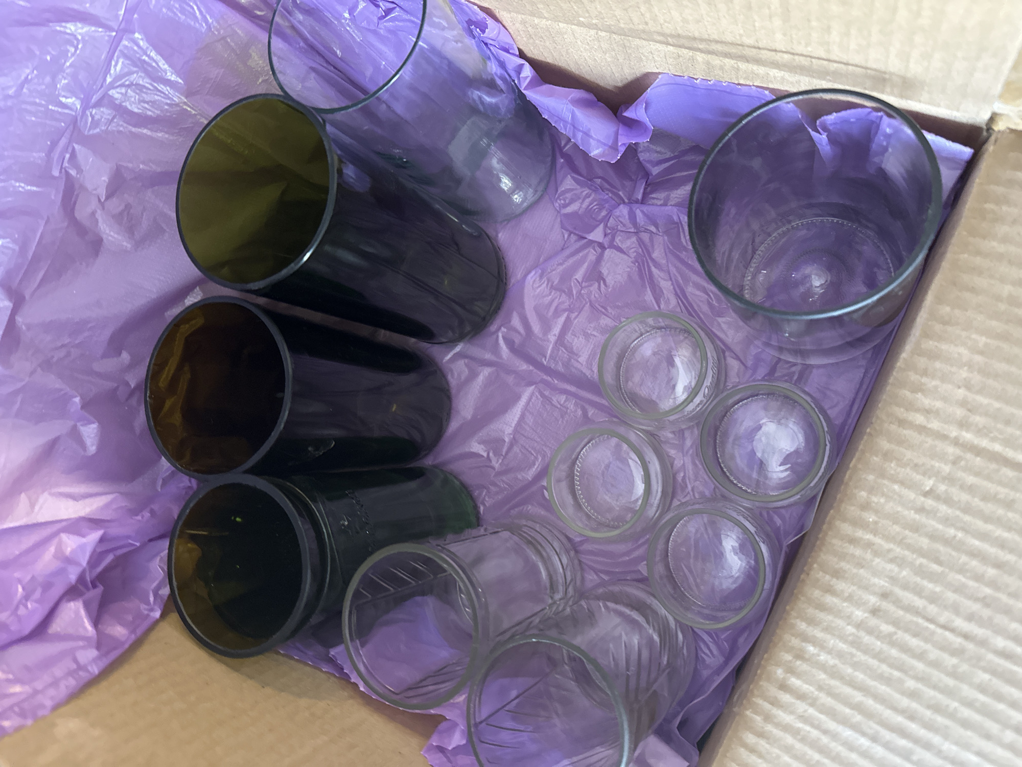 The image displays an assortment of glass containers packed inside a cardboard box, which is lined with crinkled purple tissue paper. The scene is likely indoors in Kuda, Russia.
The objects in the box consist of various types of glass items. On the left side, there are five dark, possibly brown or dark green, cylindrical glass containers, seemingly repurposed from bottle bottoms, lying horizontally. These are uniform in size and shape, suggesting they might be used as glasses or small vases.
To the right, there are numerous clear glass items. Six smaller clear glass jars are arranged in two rows of three, likely repurposed from food jars. Above these, towards the top right, a larger clear glass tumbler or jar stands upright. Another larger clear glass container is partially visible in the upper central part of the box.
The arrangement suggests the items are either being carefully packed for storage or transport, or have just been unpacked. The crinkled purple tissue paper serves as cushioning, indicating care for the fragile contents. No people are visible, and there are no specific cues regarding the time of day or weather. No discernible text is visible on any of the objects or the packaging. The image captures a still life of repurposed glass items, possibly for craft, home decor, or retail purposes.