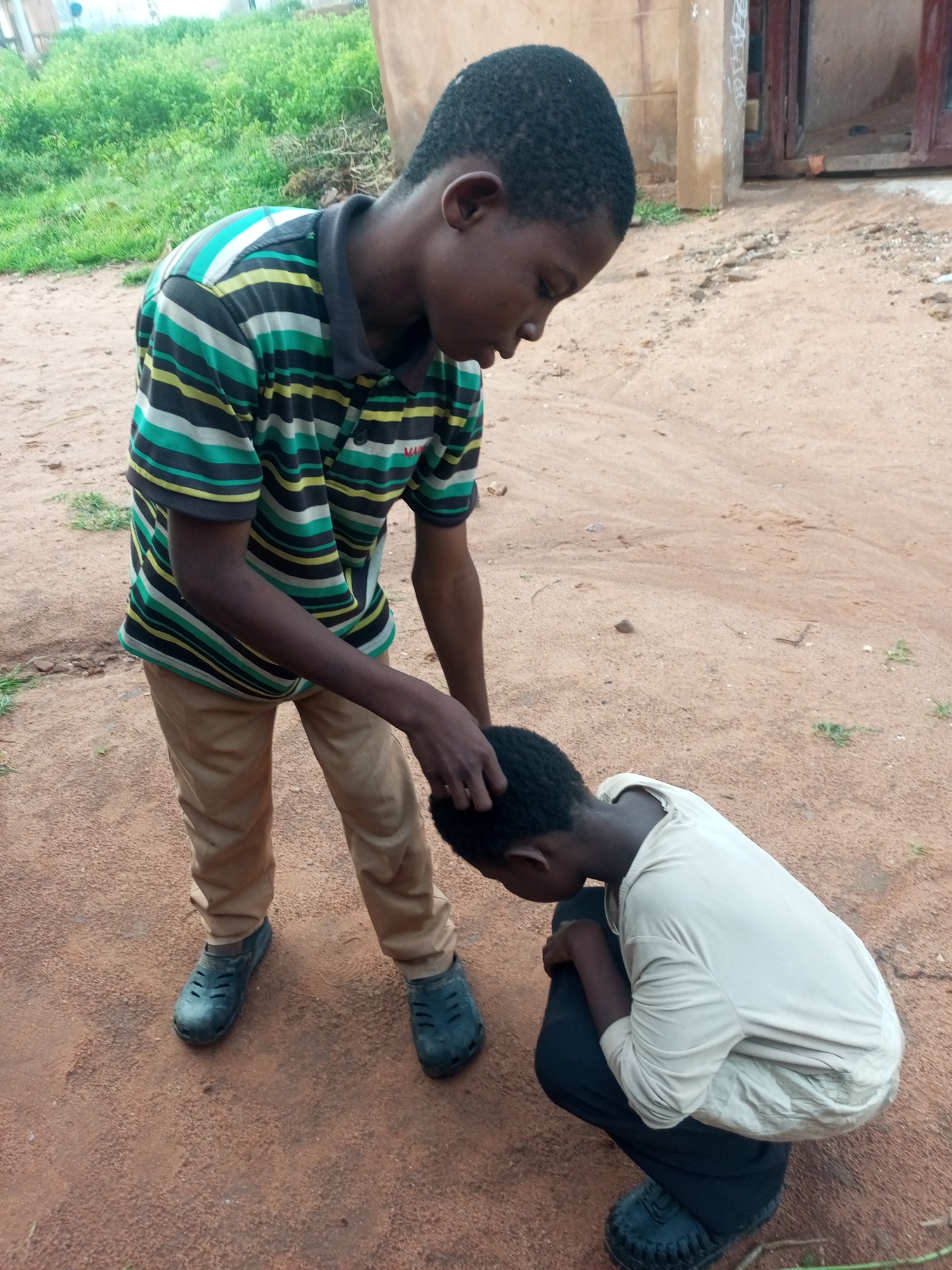 In a sandy, outdoor area with sparse green vegetation in the background, two young boys are interacting. The boy on the left, wearing a striped t-shirt and beige pants, is standing and has his hand on the head of the boy on the right. The boy on the right is kneeling and wearing a light-colored long-sleeved shirt and dark pants. The environment appears to be a rural or semi-urban setting, possibly a yard or an unpaved area within the city of Tempure, Nigeria. A beige building structure is partially visible in the background.