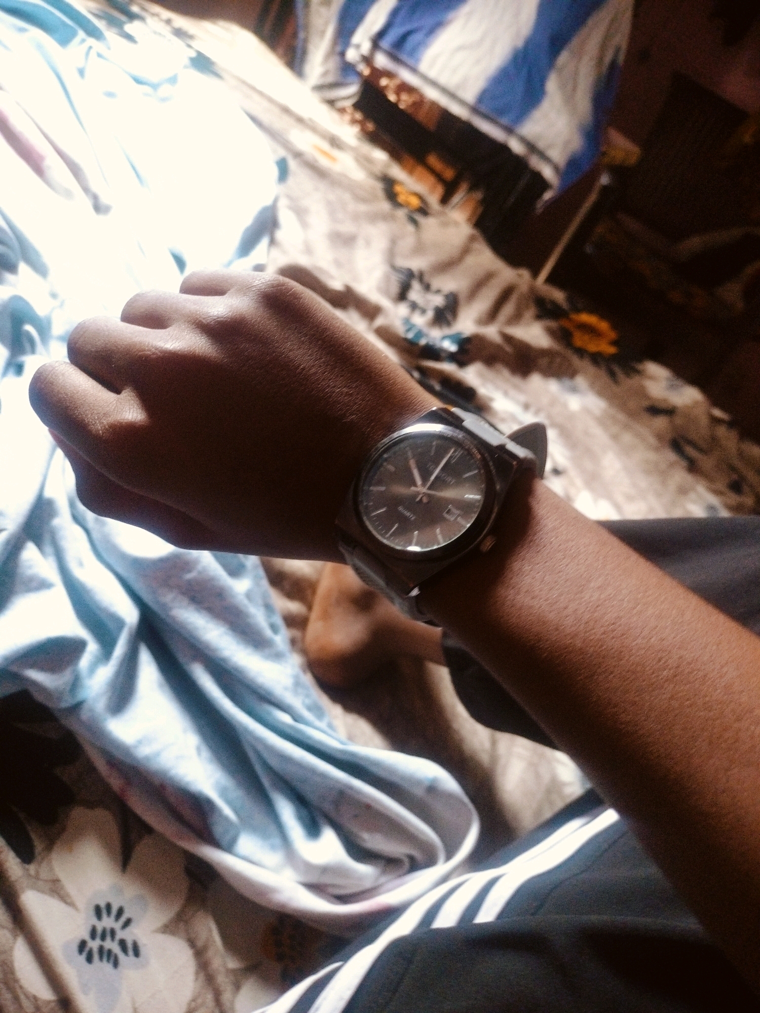 A close-up, overhead shot shows a person's left arm and hand wearing a dark gray watch with a silver face, resting on a bed with patterned sheets. The sheets are a mix of floral and striped patterns in light blue, white, and brown hues. The person's skin appears to be dark brown. The image is dimly lit with some bright spots, suggesting it was taken indoors, possibly in a bedroom in Un, India. The scene is static and shows no interaction or action.