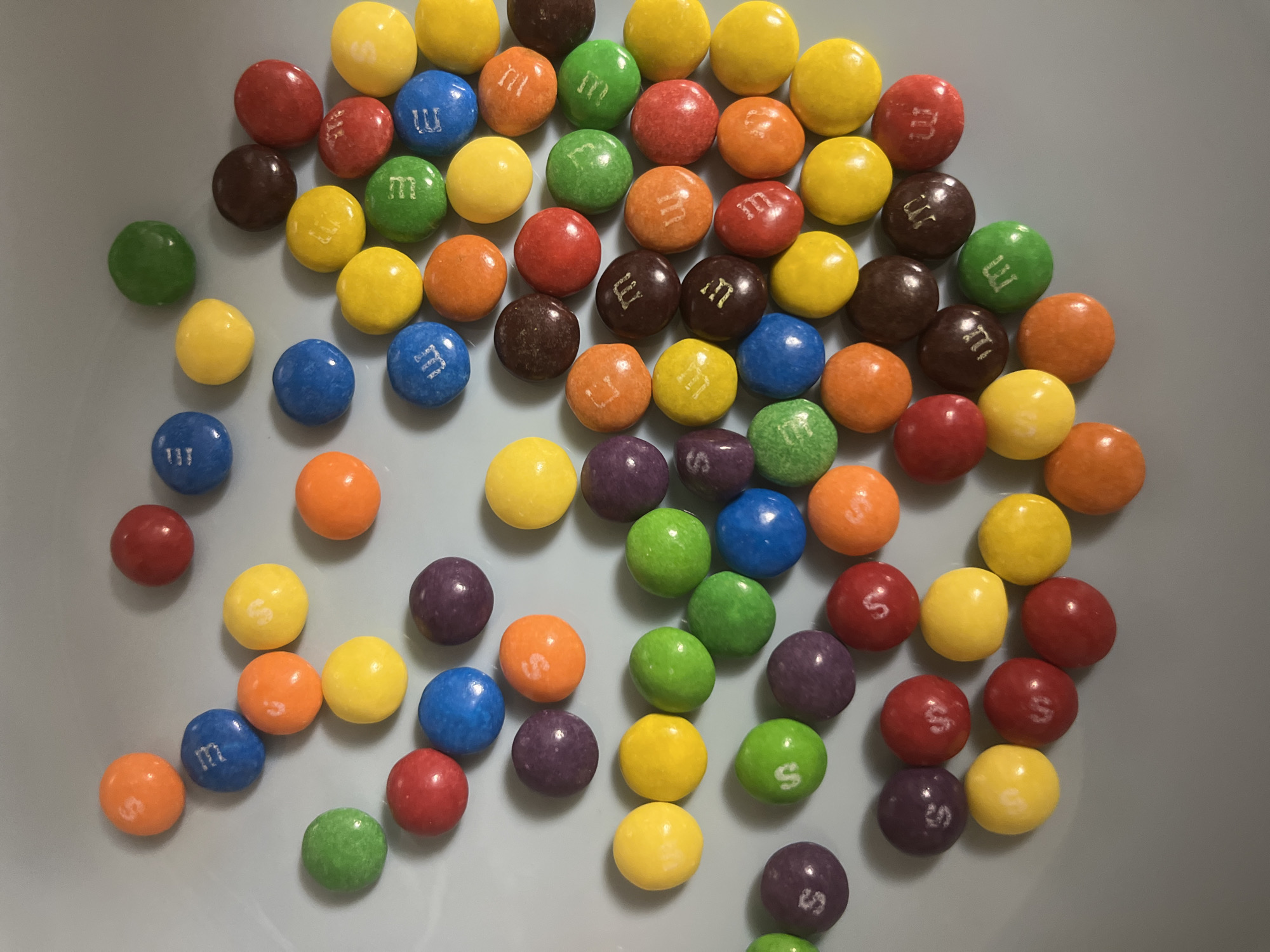 A slightly overhead shot displays a pile of M&M candies scattered on a light grey surface. The candies are in various colors including yellow, red, blue, orange, green, brown, and purple. The letter "M" is printed in white on several of the candies. The edges of a plate or bowl are visible. The location is unknown.