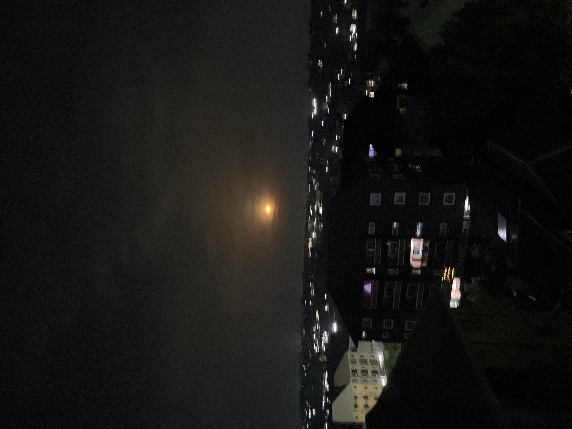 The image presents a night view in Nkpor, Nigeria. The right side of the frame displays tall buildings illuminated by numerous lights. The left side is dominated by a dark sky with an indistinct, soft glow suggestive of a hazy moon or light pollution. A few trees are barely visible on the right side.