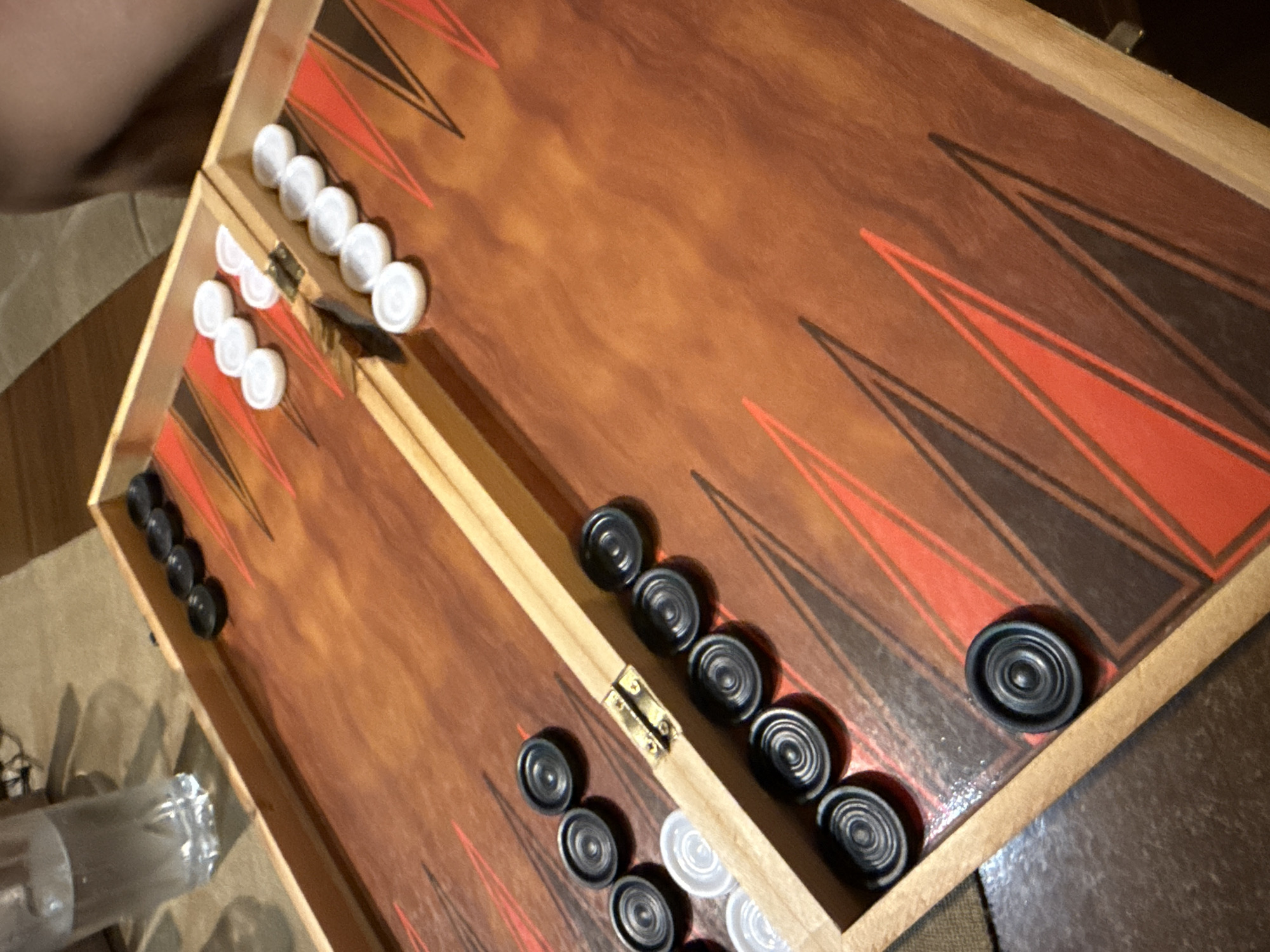 The image shows a backgammon board with checkers. The board is made of wood with a reddish-brown finish and triangular markers in red and black. On the left side of the board, there are black checkers arranged in two groups, one of five checkers and another of four checkers. Near the hinge, there are also two white checkers. On the right side of the board, there are white checkers arranged in three groups, one of three checkers, another of two checkers, and a single checker. There are also several black checkers on the right side of the board. A glass of water is visible in the upper left corner. The scene appears to be indoors, possibly on a table. The specific location in Blagoevgrad, Bulgaria, cannot be determined from the image.