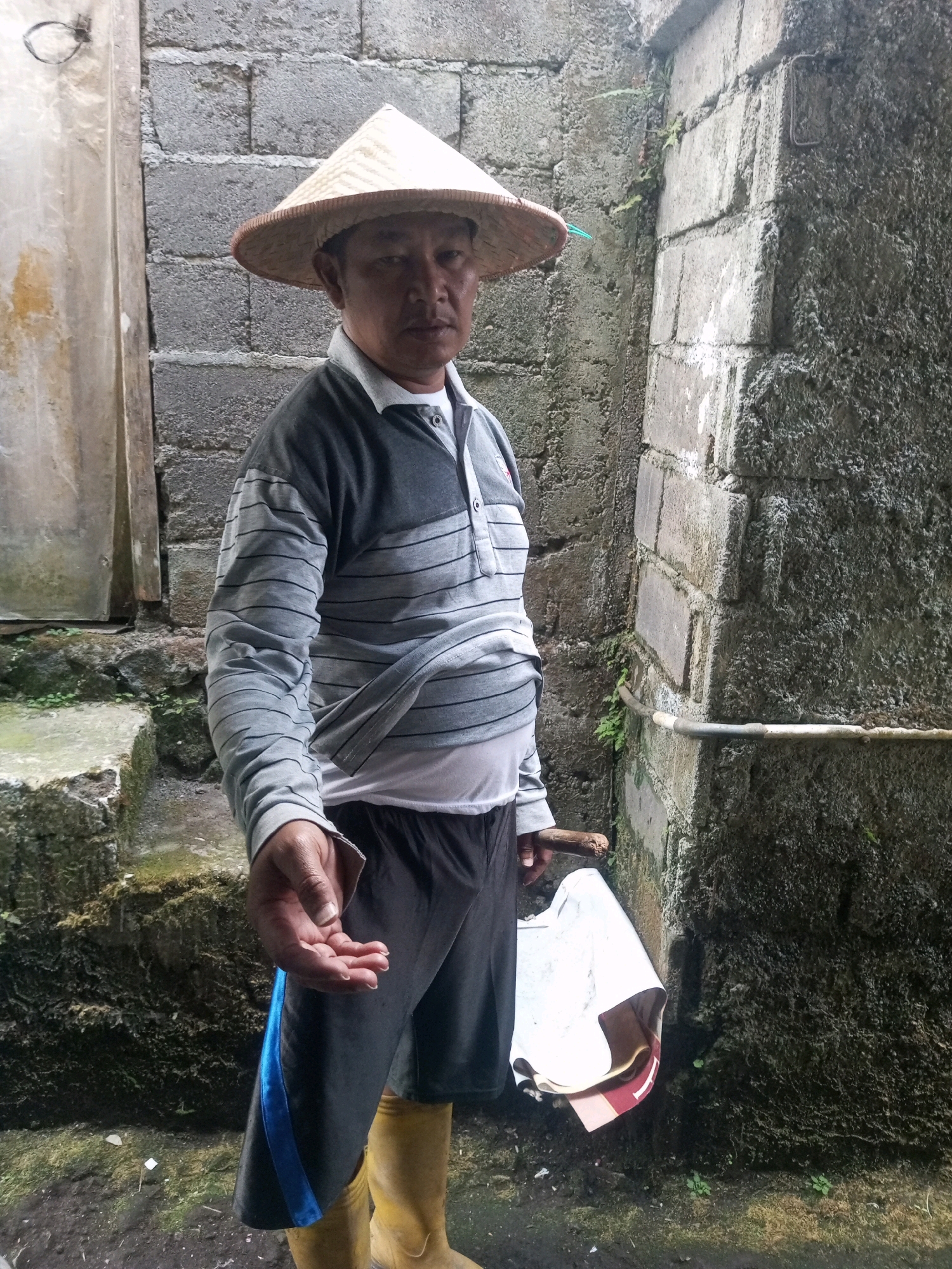 An Indonesian man stands in front of a rough concrete wall, wearing a conical straw hat, a grey and white horizontally striped long-sleeved shirt, dark shorts with a blue stripe, and yellow rubber boots. He holds a white cloth bag in his right hand and a wooden stick in his left hand. The background consists of a textured concrete wall and some green vegetation. The ground is muddy. The image appears to have been taken outdoors in what may be a rural or semi-urban area in Andongsili, Indonesia, given the rustic construction and vegetation.