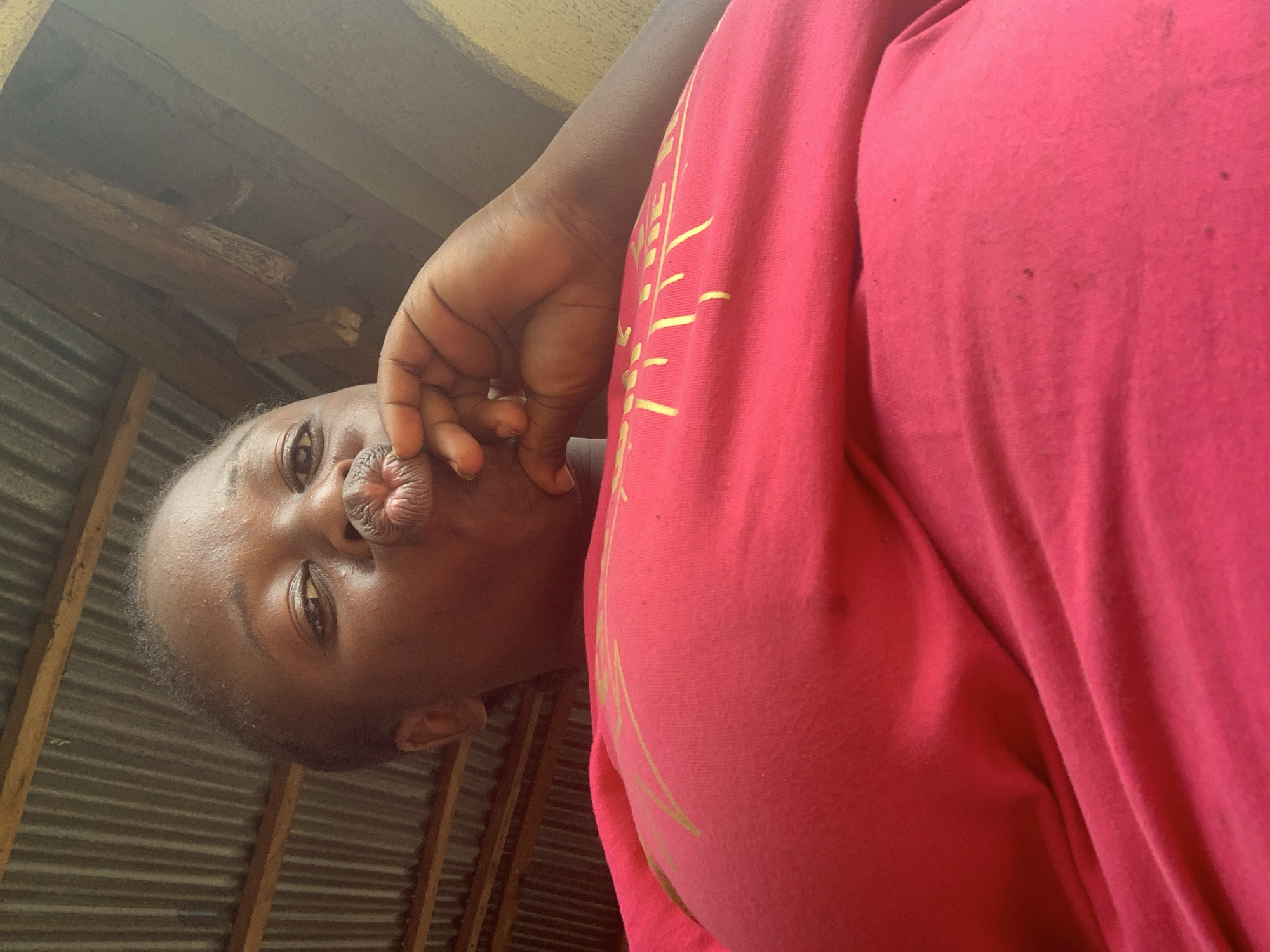 The image displays a close-up, low-angle portrait of a young Black individual, presumed female, located indoors within the city of Akwanga, Nigeria. The individual is positioned centrally, directly facing the camera with open eyes and puckered lips, forming a "kiss" expression. Her left hand is raised, with fingers resting against her chin and lower lip. She is wearing a vibrant pink short-sleeved top with a faint yellow, possibly graphic, design across the upper chest. The background elements include a corrugated metal roof structure, visible on the upper left, supported by prominent wooden beams and rafters extending across the top and right side of the frame. A light-colored wall surface is partially visible on the far right.